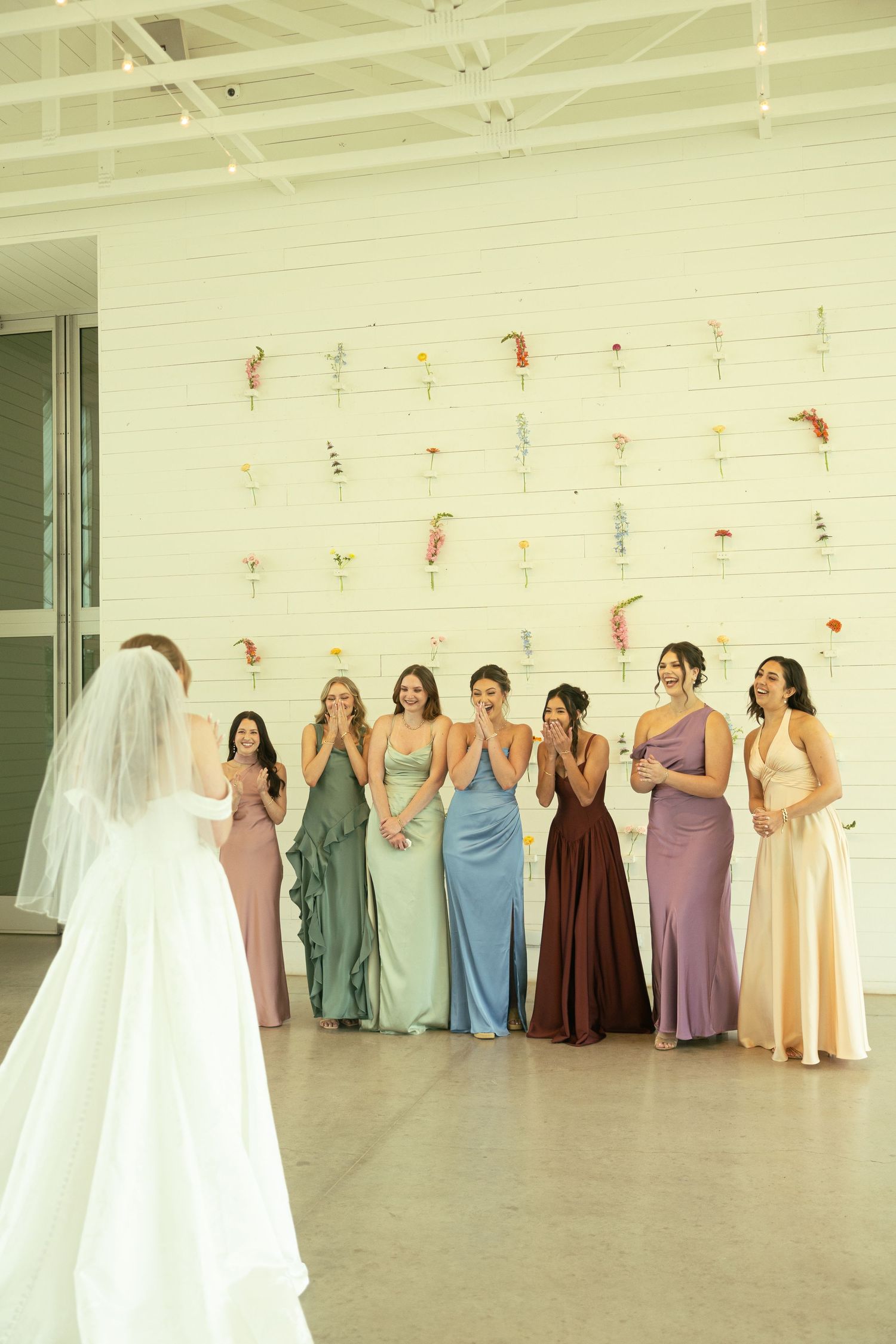 Bride reveals wedding dress to bridesmaids in colorful gowns against a floral wall backdrop.