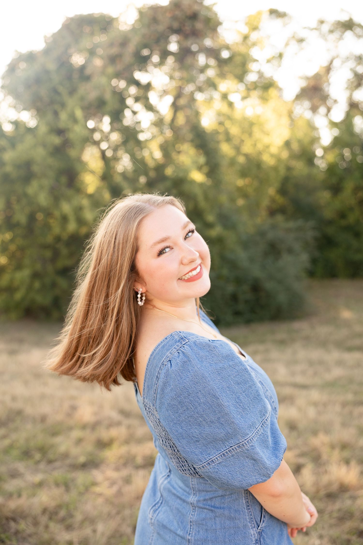 Person in denim dress smiling over shoulder in sunlit outdoor setting.