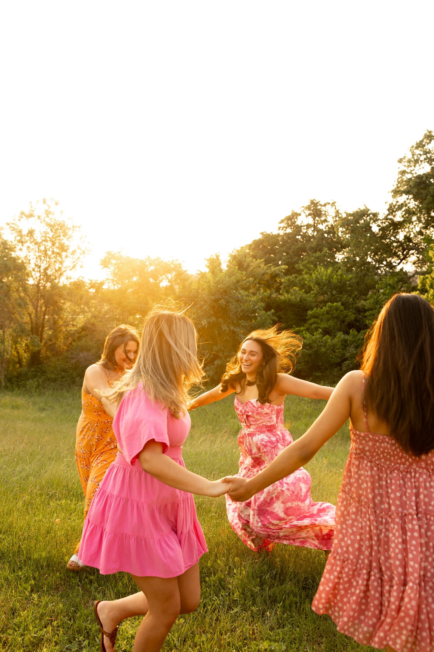 Friends in flowing dresses dance together in a sunlit meadow at dusk.