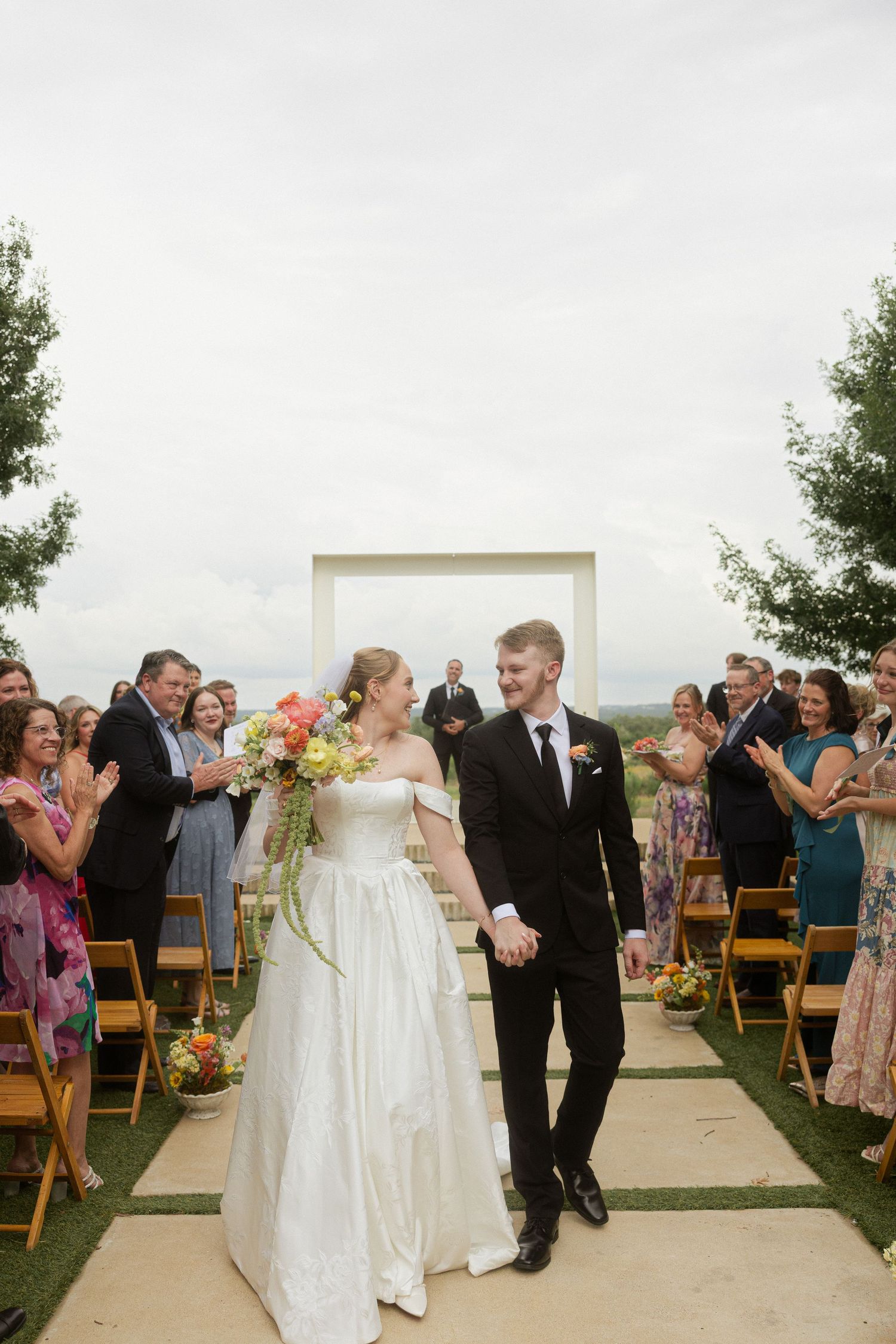 Bride and groom walk down outdoor wedding aisle, guests applauding, white arch backdrop, bride holding colorful bouquet.