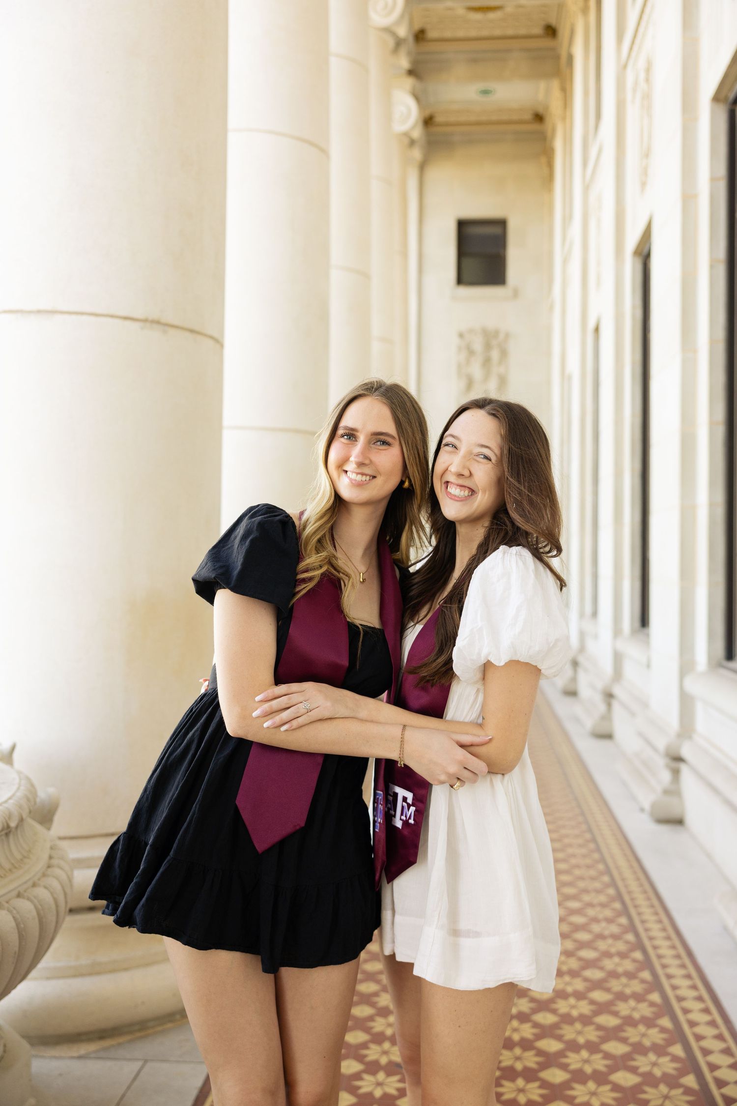 Two graduates in black and white dresses with maroon stoles embrace in columned hallway.