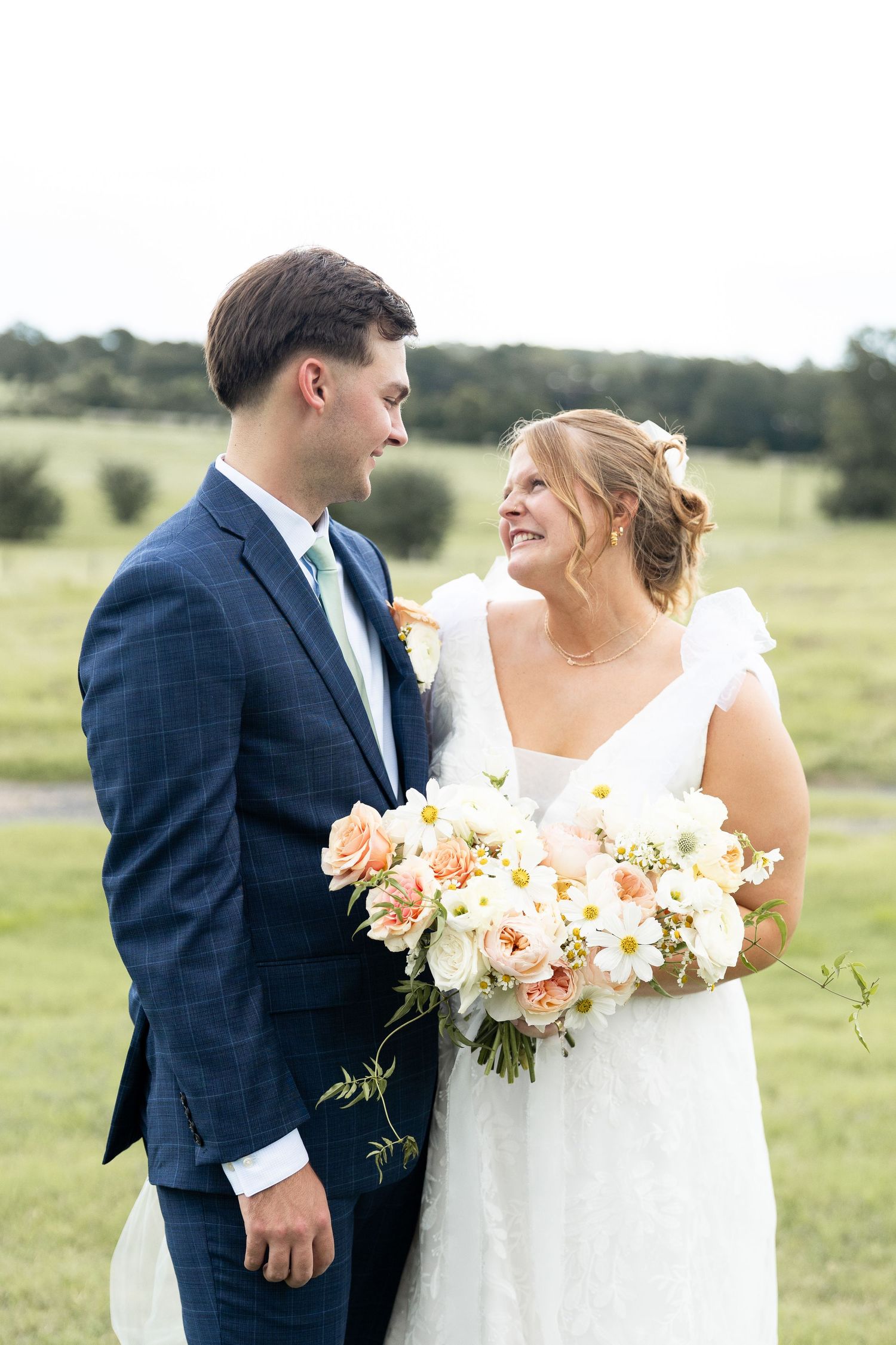 A romantic outdoor portrait with soft peach and white bouquet against a pastoral landscape.