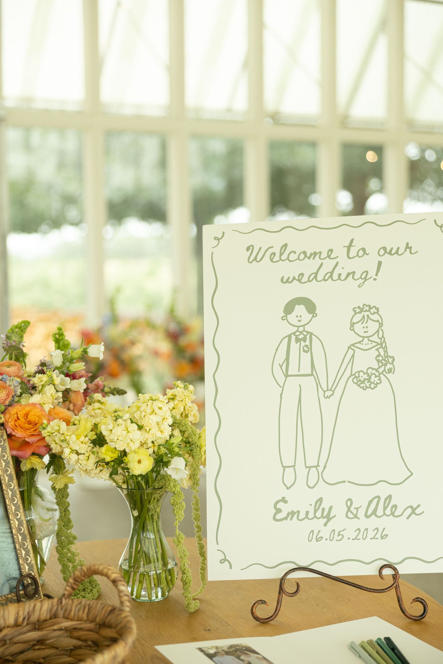 Wedding welcome sign for Emily and Alex dated 06.05.2026 with illustrated couple beside fresh floral arrangement.