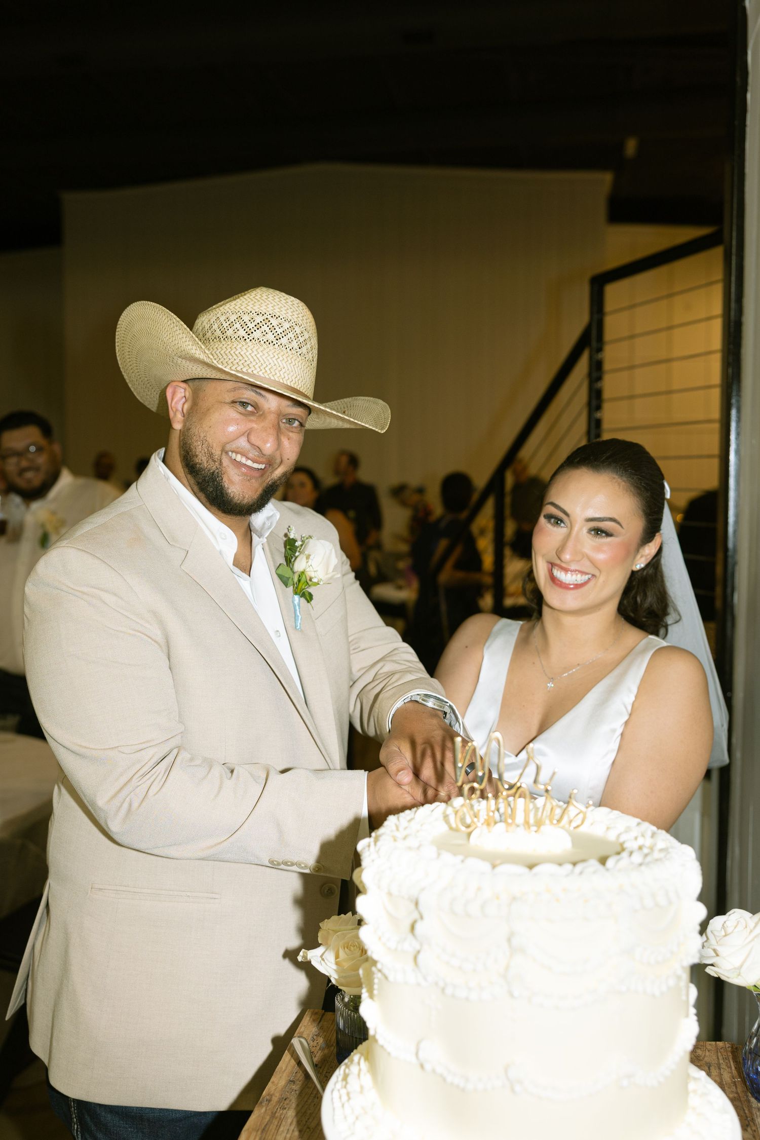 A couple in white western attire share a joyful moment cutting their wedding cake together.