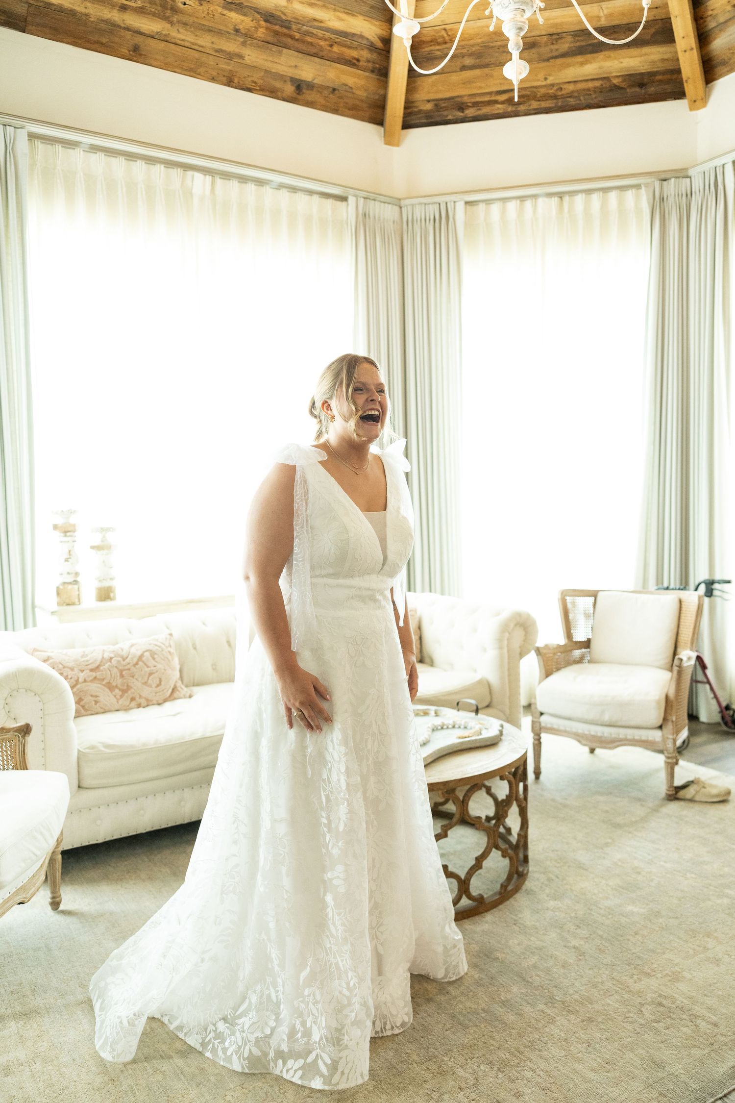 An elegant white lace wedding dress is showcased in a sunlit room with rustic wooden ceiling beams and white furniture.
