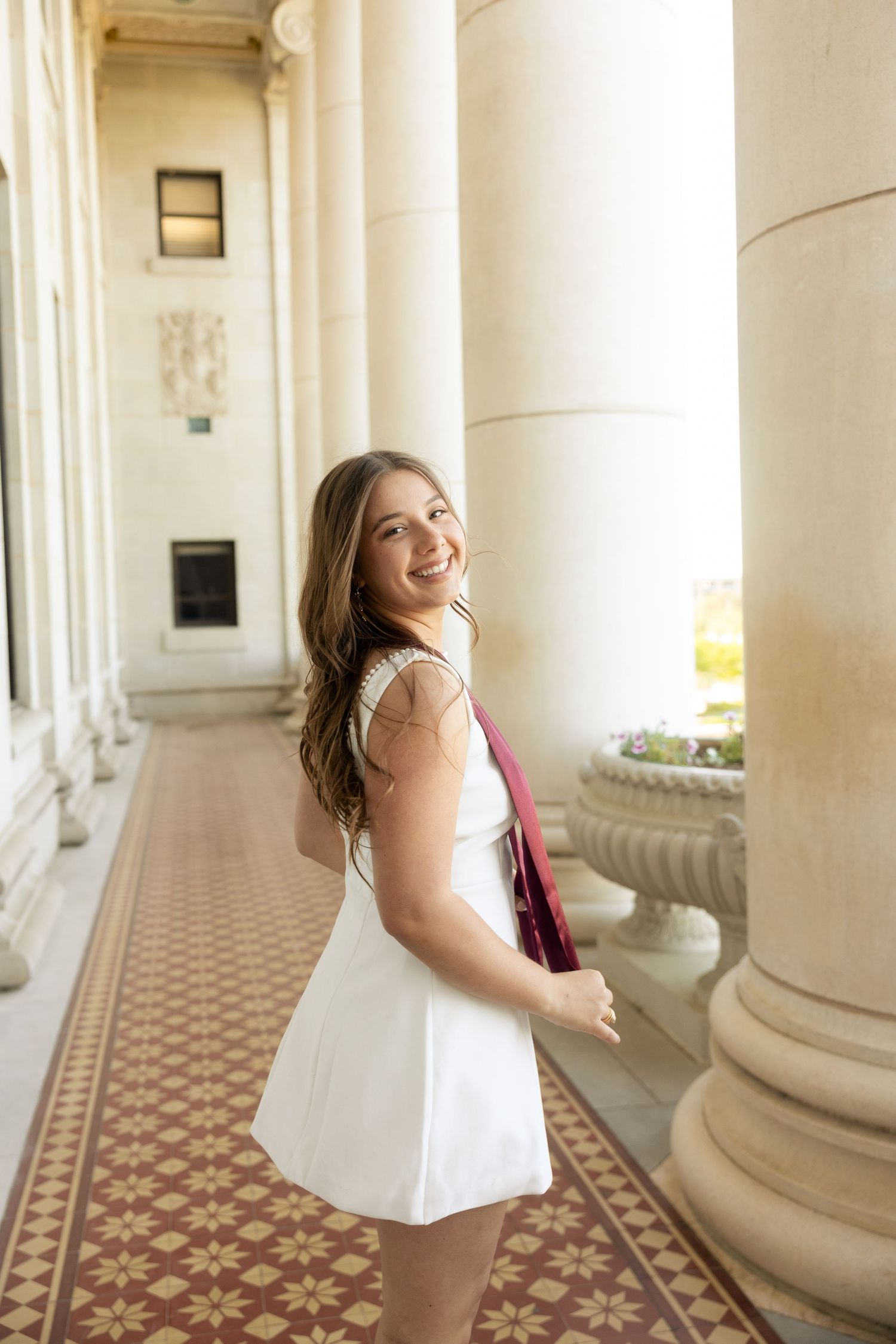 Graduate in white dress poses joyfully between classical columns.