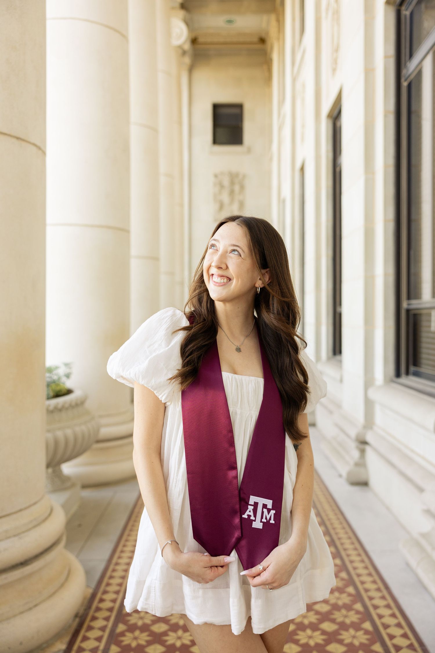 Graduate in white dress with maroon Texas A&M stole stands in columned hallway with patterned floor.