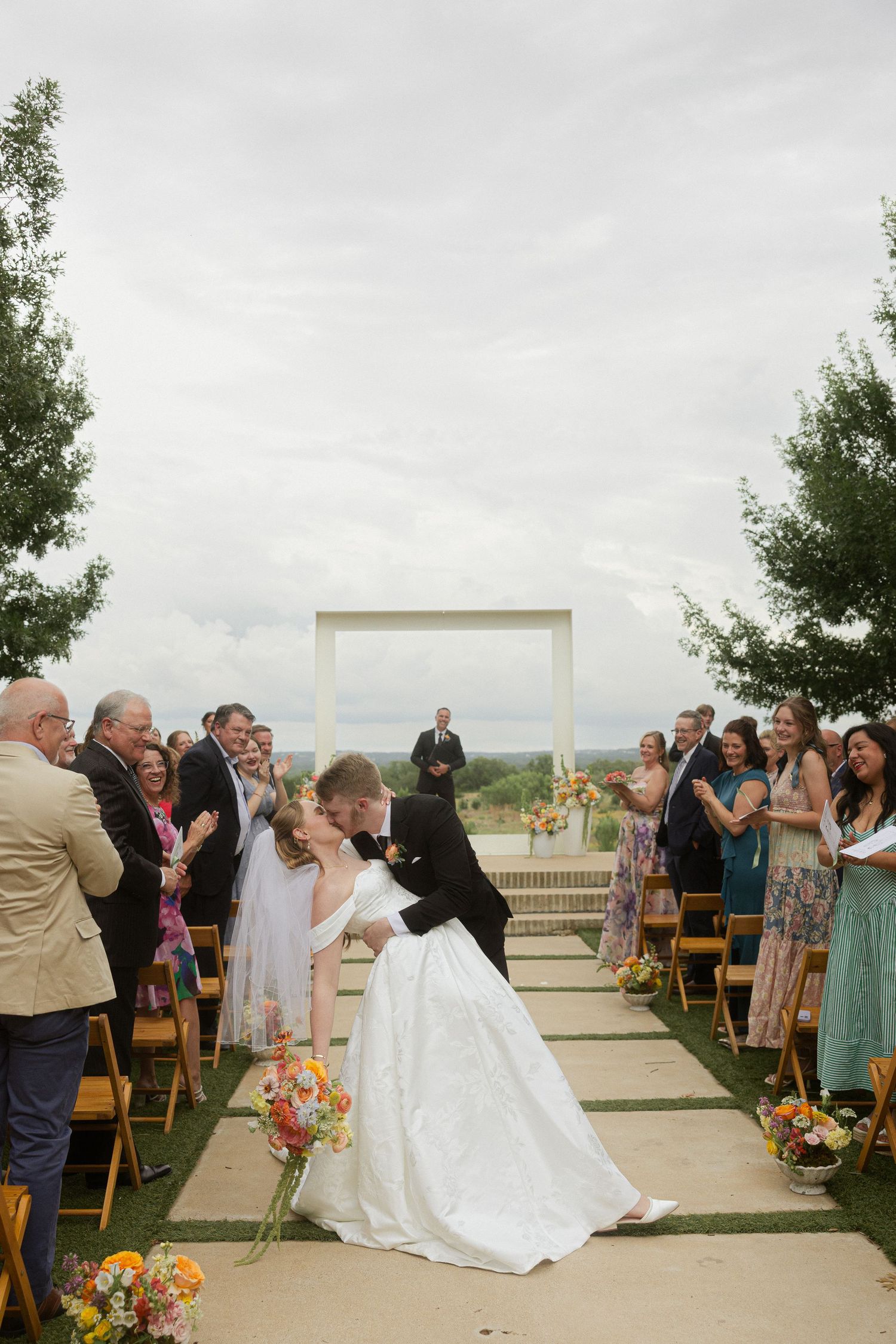 Bride and groom share romantic dip kiss at outdoor wedding ceremony aisle with guests applauding.