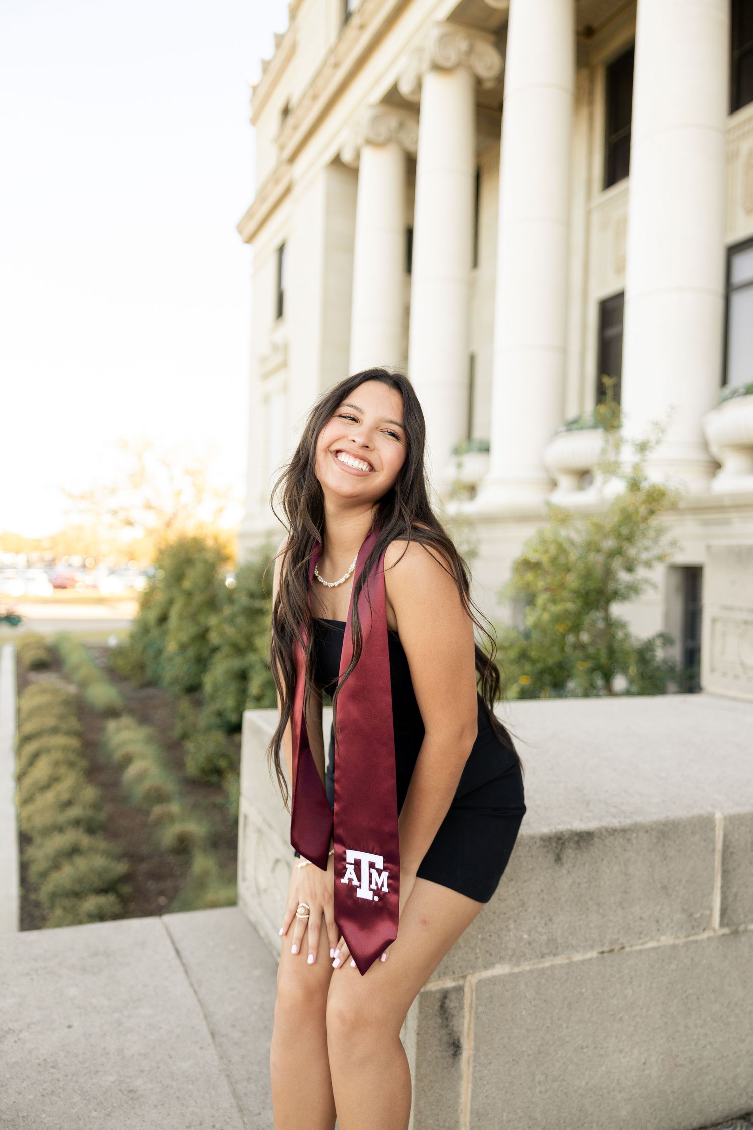 Joyful graduate in black dress and maroon Texas A&M stole poses near columned building.
