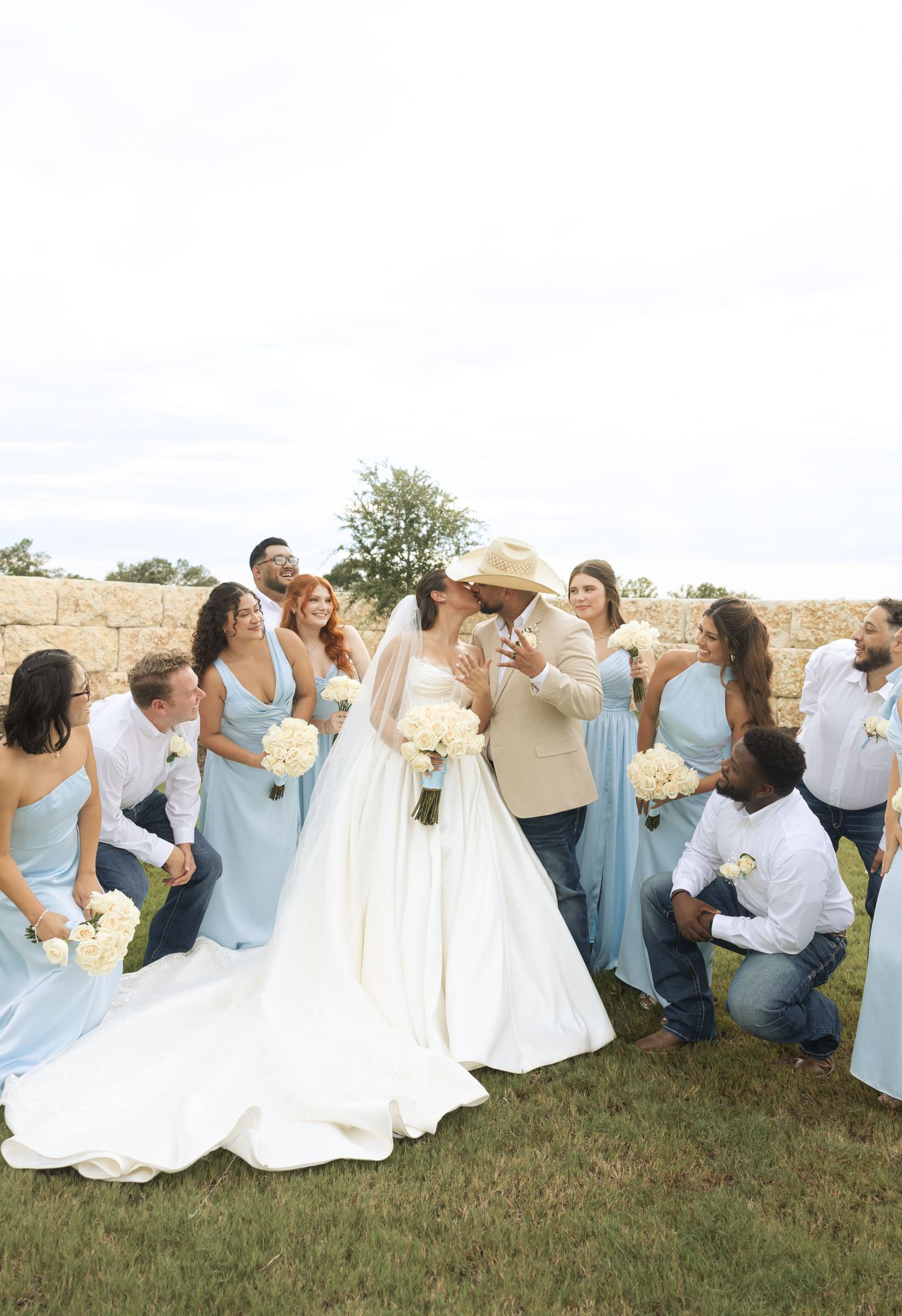 Wedding party in light blue dresses and white gowns pose playfully on grassy lawn against stone wall backdrop.