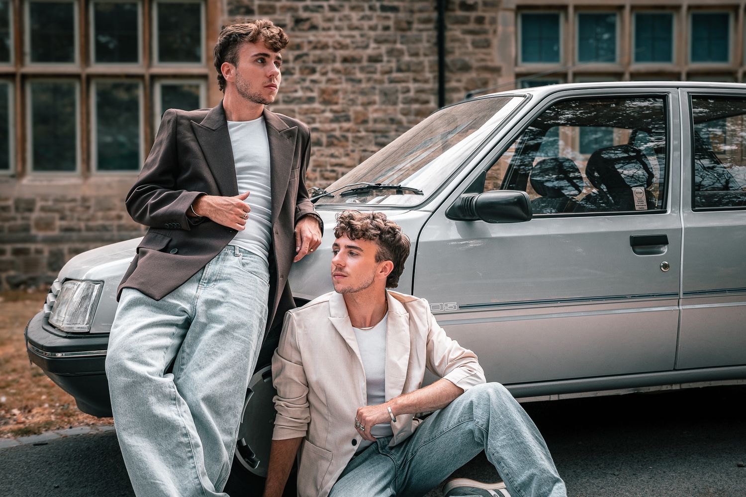 Two stylish young men posing near a silver car, wearing blazers and jeans in a urban setting.
