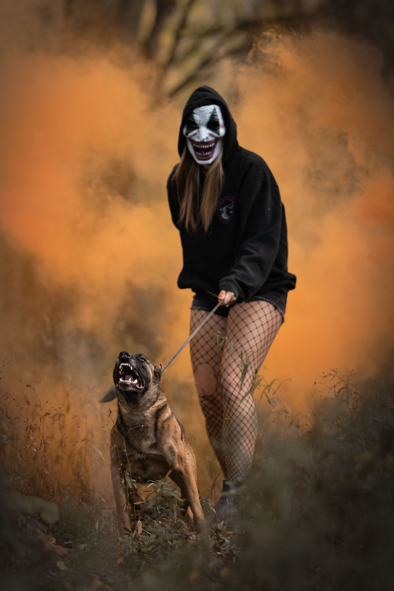 A person in clown makeup and fishnet stockings stands with a fierce dog amid orange smoke in a dark atmospheric setting.