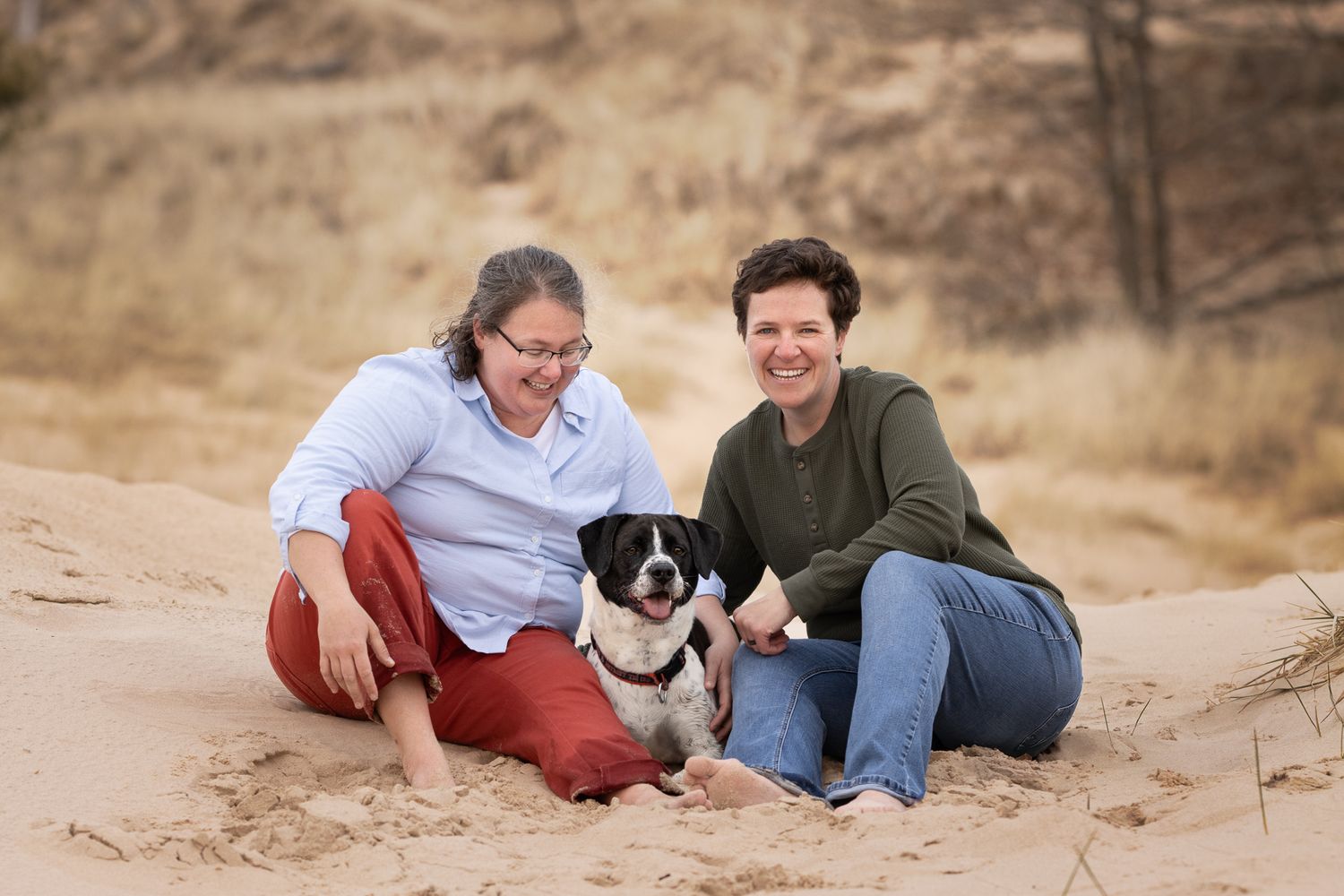 A couple sits on sandy dunes with their black and white border collie puppy during a casual outdoor photo session.