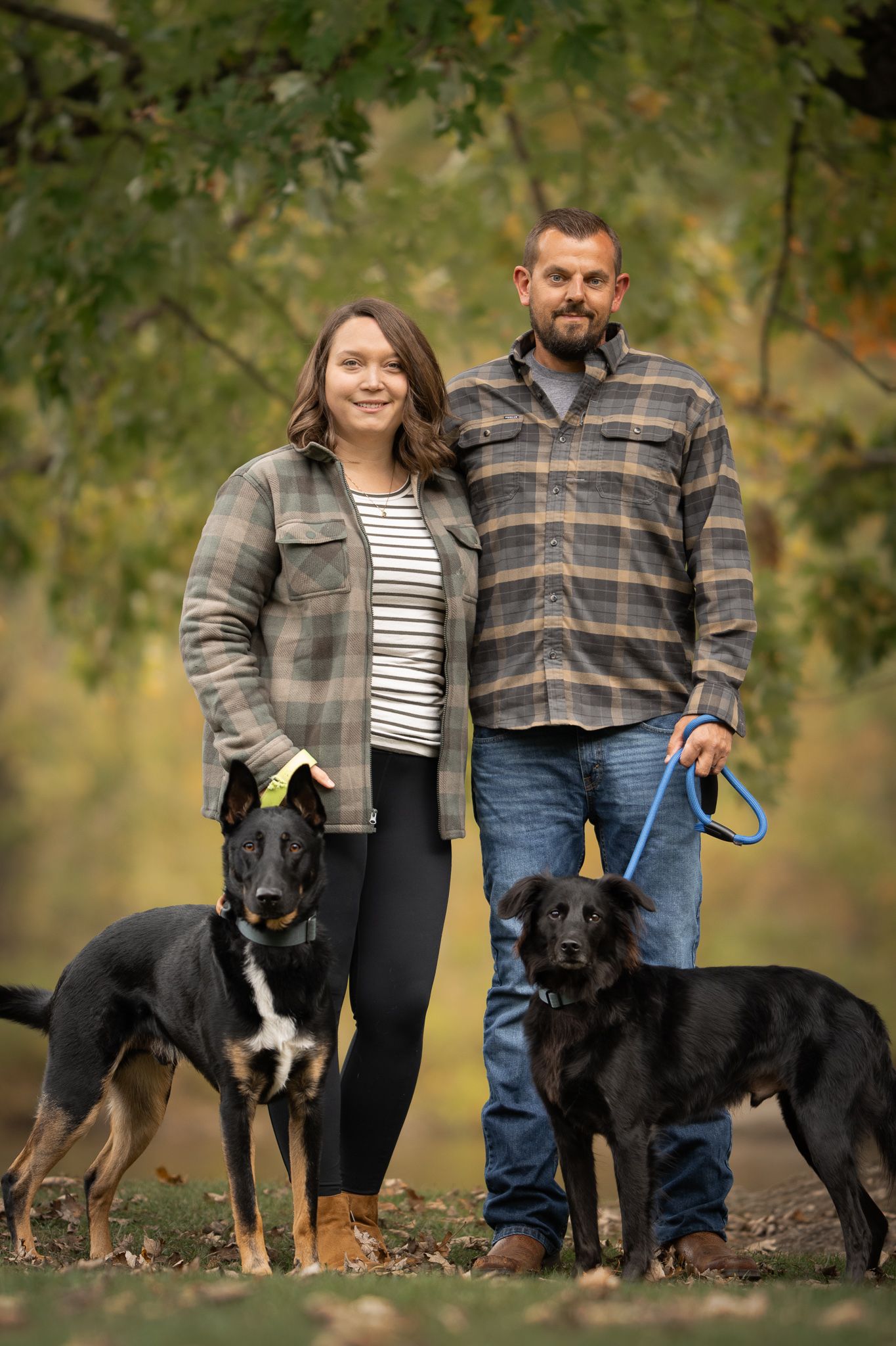 A couple poses outdoors with their three dogs including a black and white border collie mix and two dark-colored dogs.