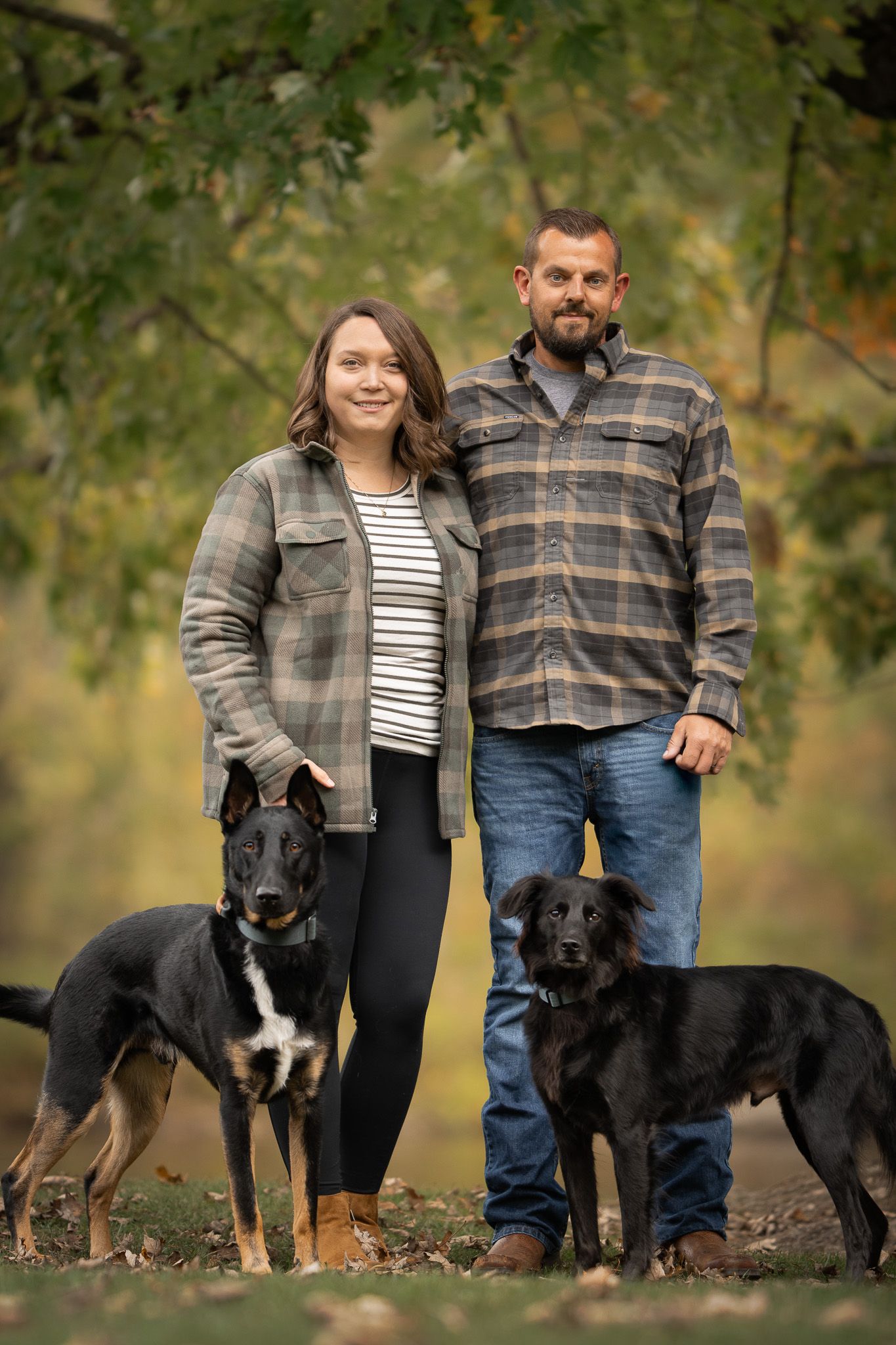 Two black dogs pose with their owners in a fall setting with trees and leaves in the background.