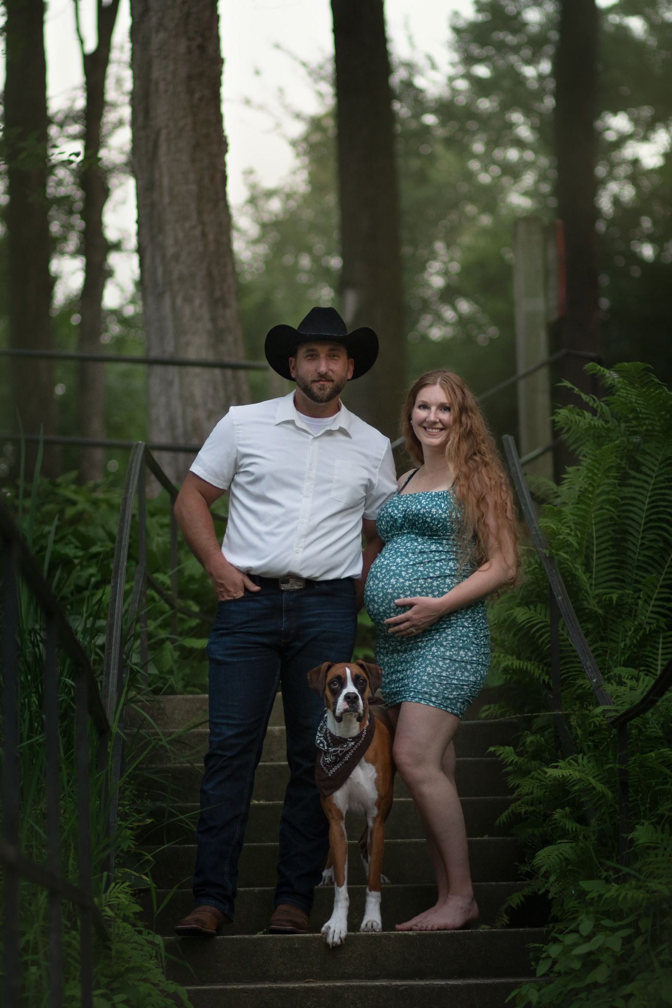 Couple poses on outdoor steps with their boxer dog surrounded by lush green ferns in a forest setting.