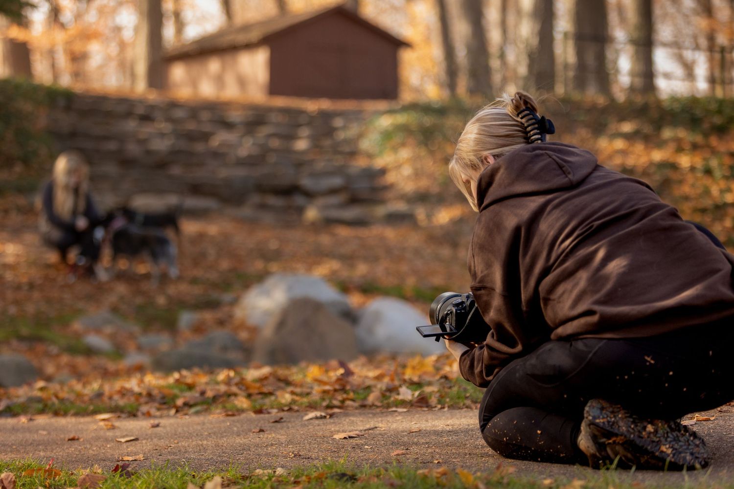 A photographer crouches low to capture photos in a park with autumn leaves on the ground during golden hour.