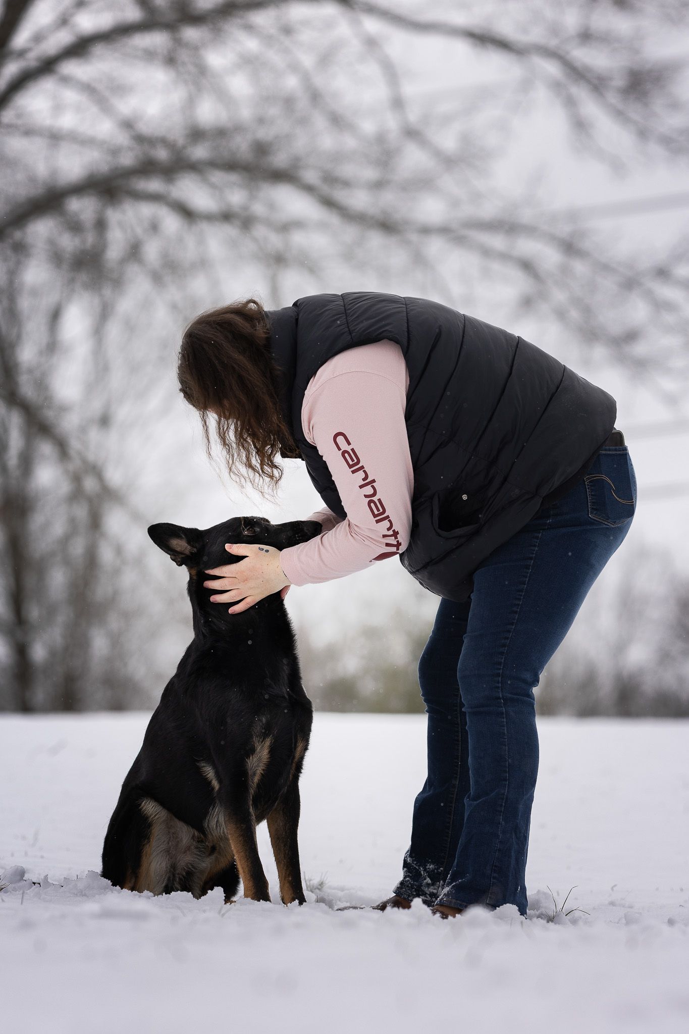 A person in a black jacket bends down to pet a black and tan dog in a snowy winter landscape.