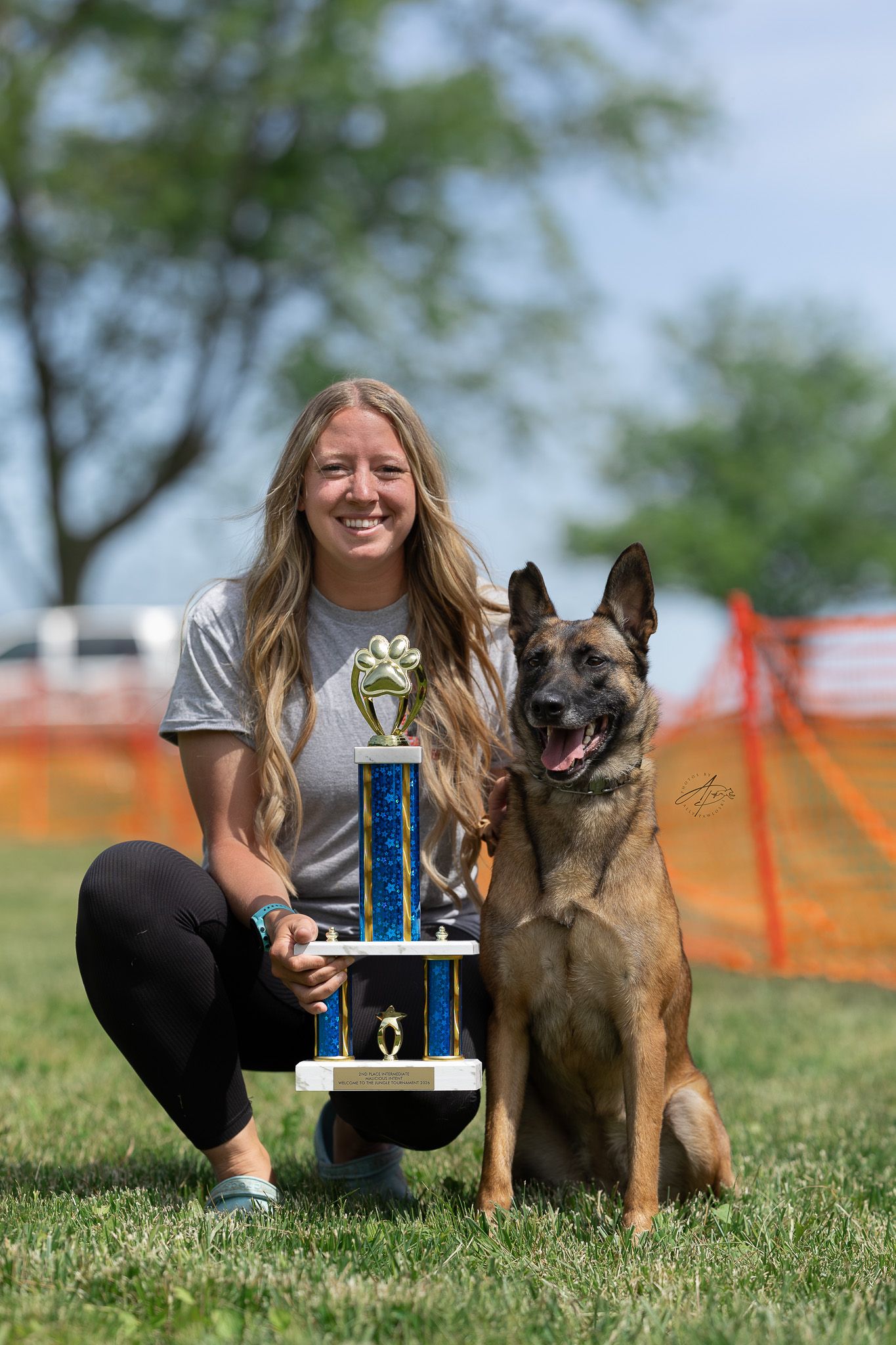 Smiling woman kneeling on grass with a Belgian Malinois dog and a blue trophy at an outdoor dog competition.