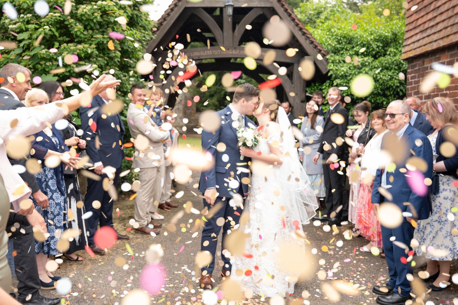 Wedding guests celebrate with confetti and bubbles outside a rustic brick building on a sunny day.