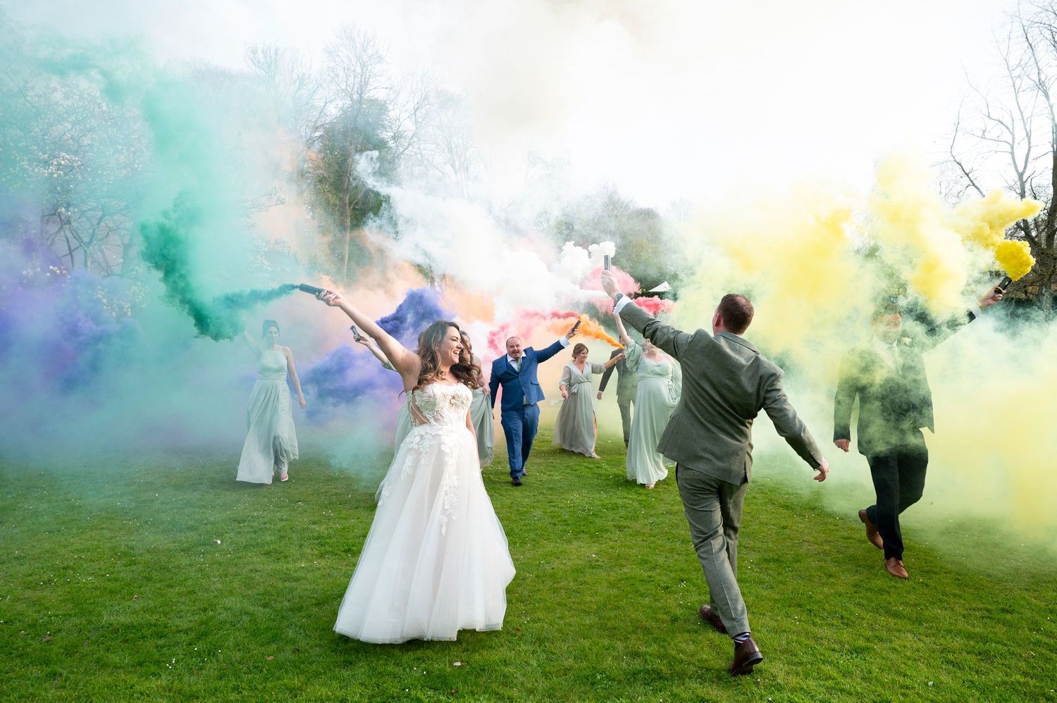 Wedding party celebrates with colourful smoke bombs on a grassy lawn. yellow, red, and purple smoke fills the scene.
