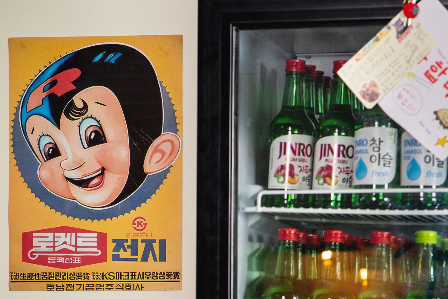 Vintage Korean cartoon poster beside a refrigerator stocked with Jinro soju bottles and colorful drinks.