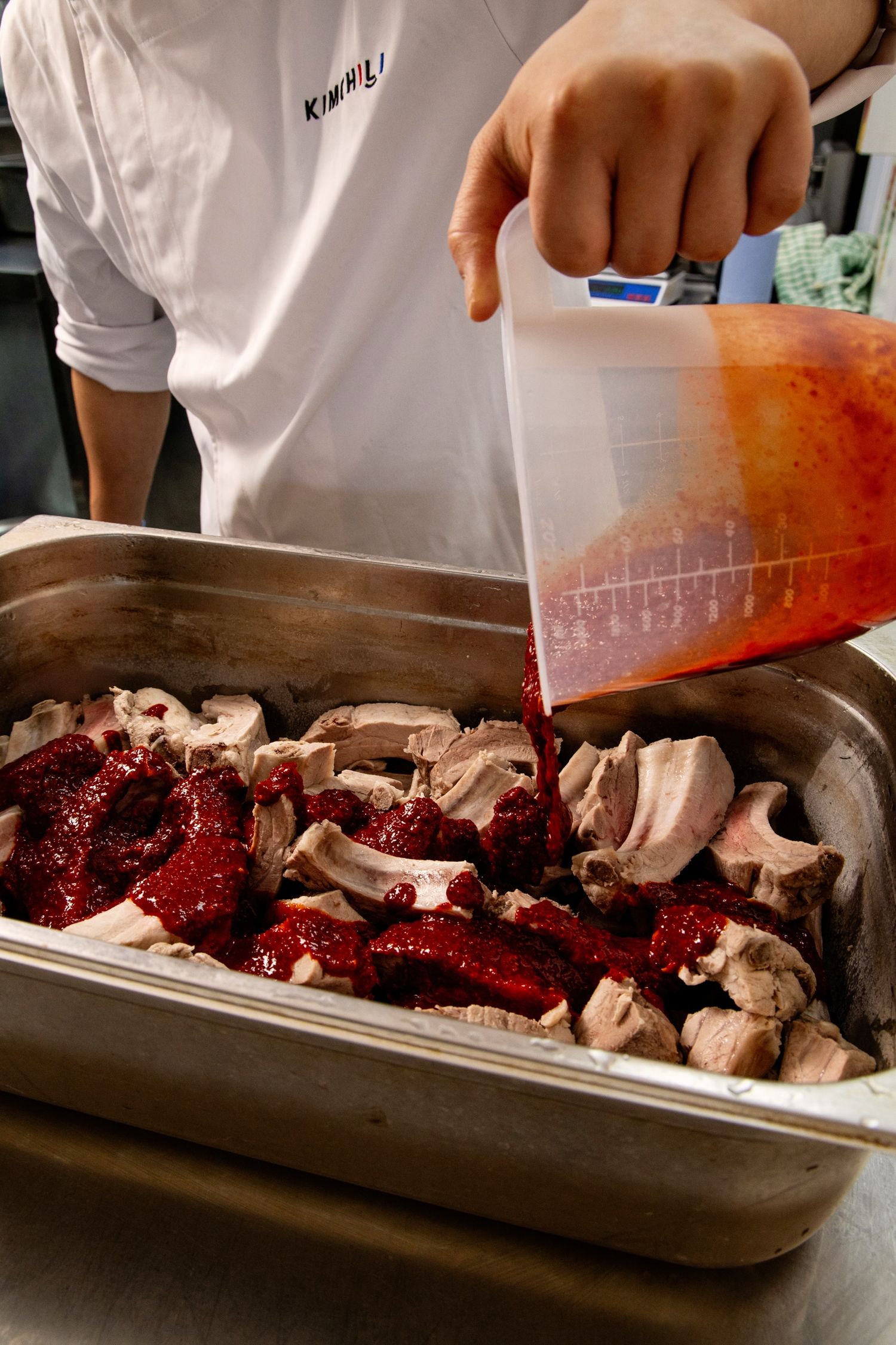 Chef pouring red marinade sauce over raw meat pieces in a stainless steel tray.