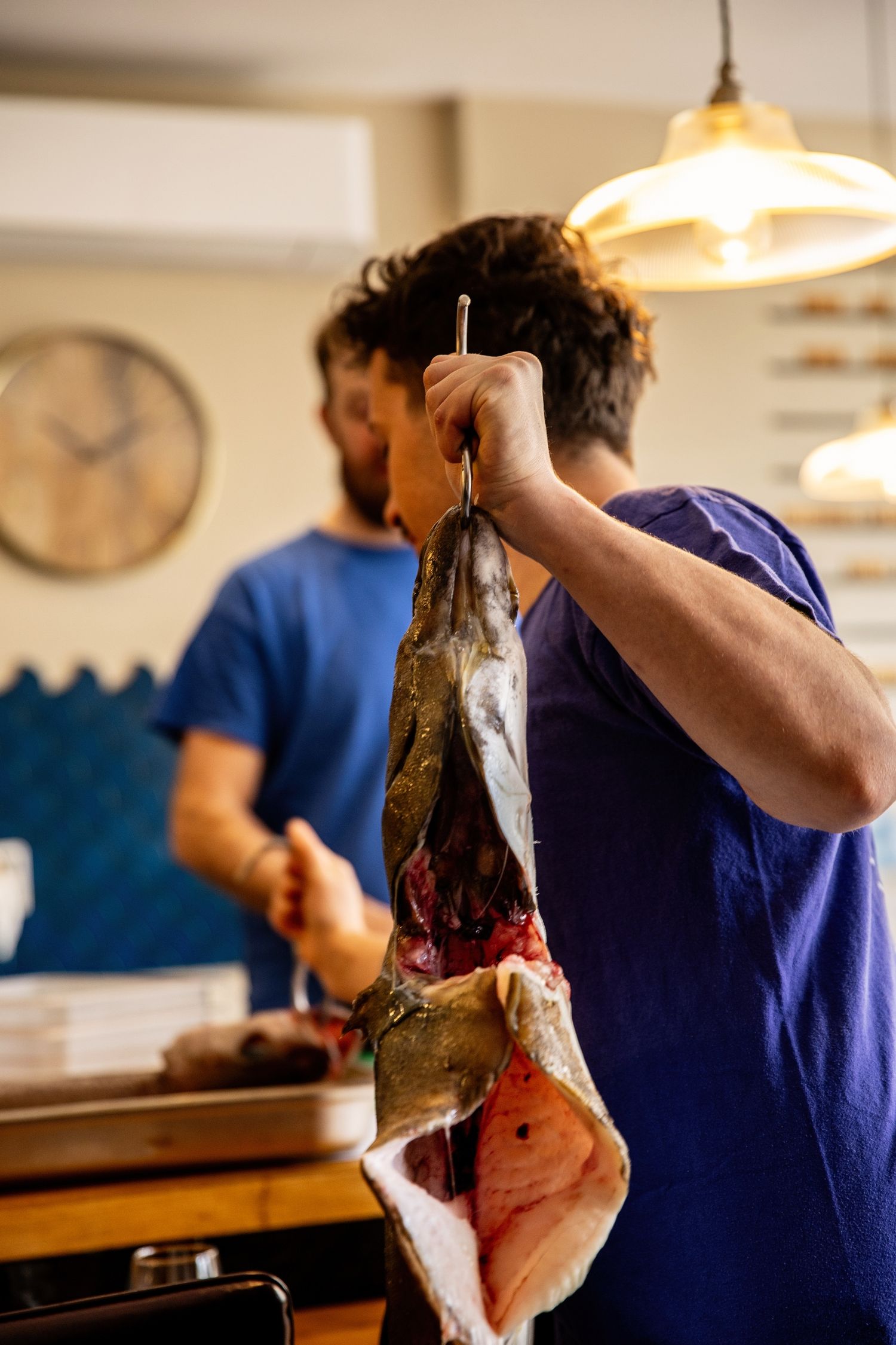 Someone in a blue shirt holds up a freshly caught fish in a kitchen with a pendant light and clock visible in background.