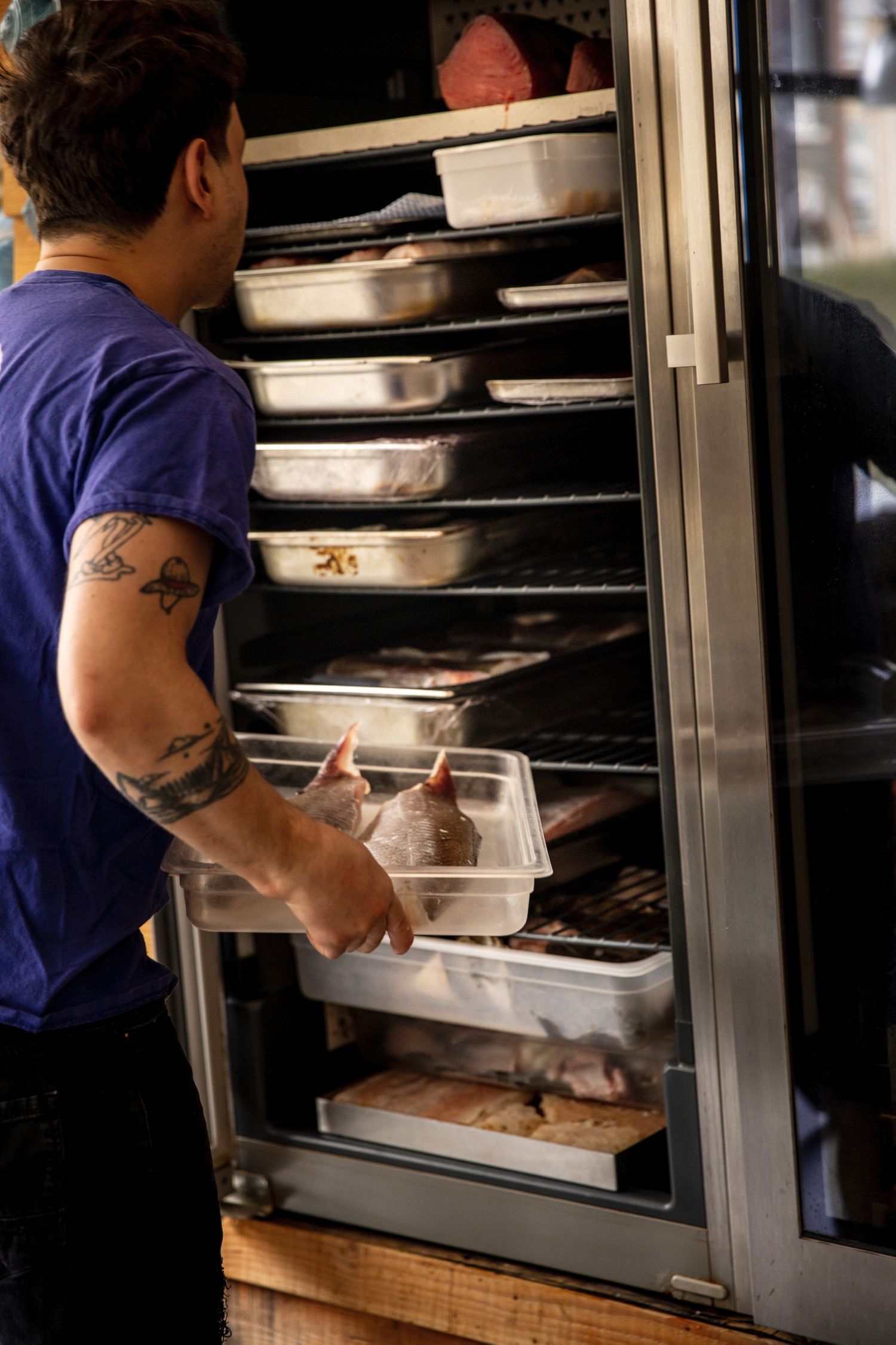 Restaurant worker with tattoo on arm placing food containers into a large industrial refrigerator with multiple shelves.