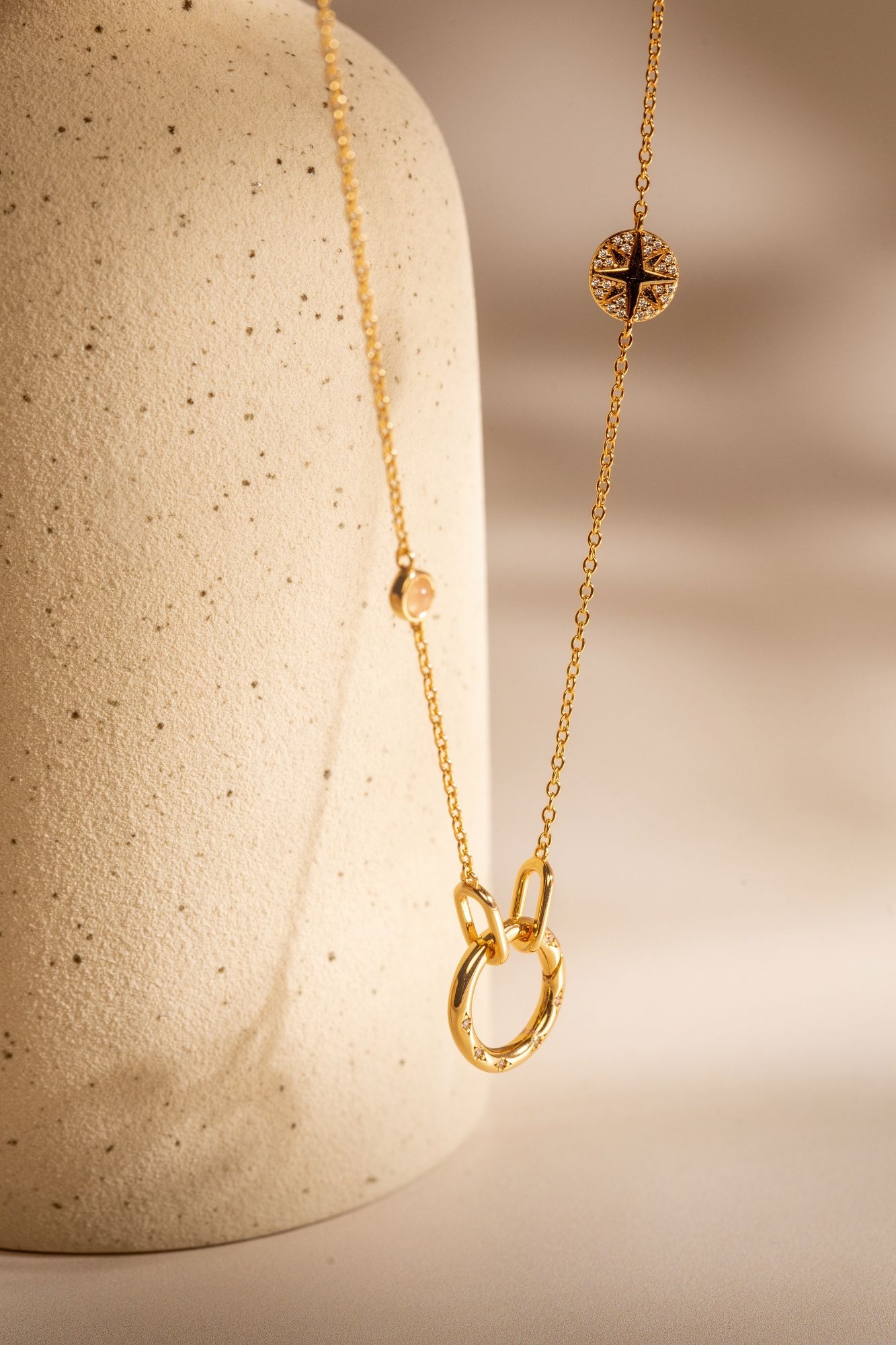 Delicate gold necklace with compass charm and circular pendant displayed against textured white surface in warm lighting.