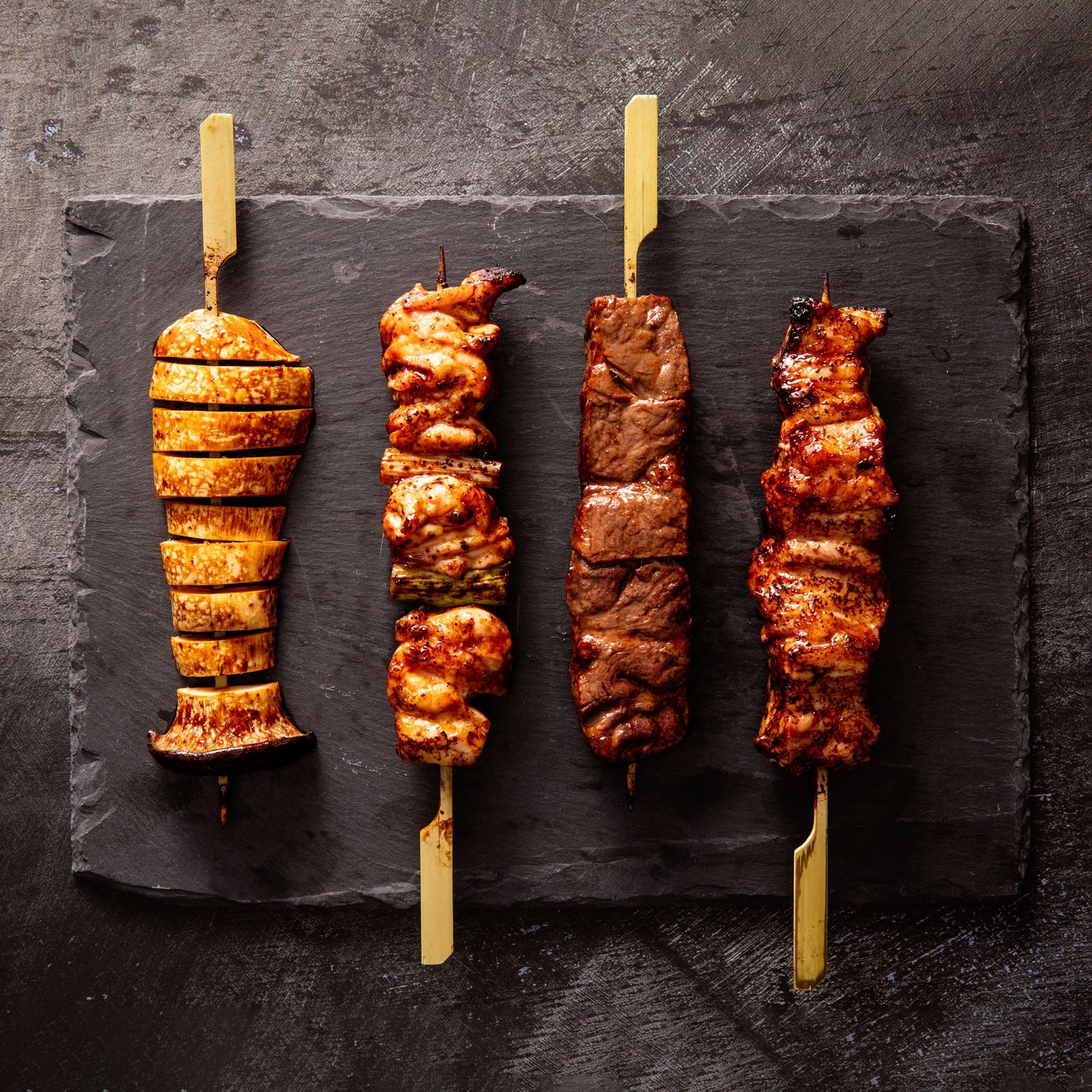 Four grilled skewers displayed on black slate surface.