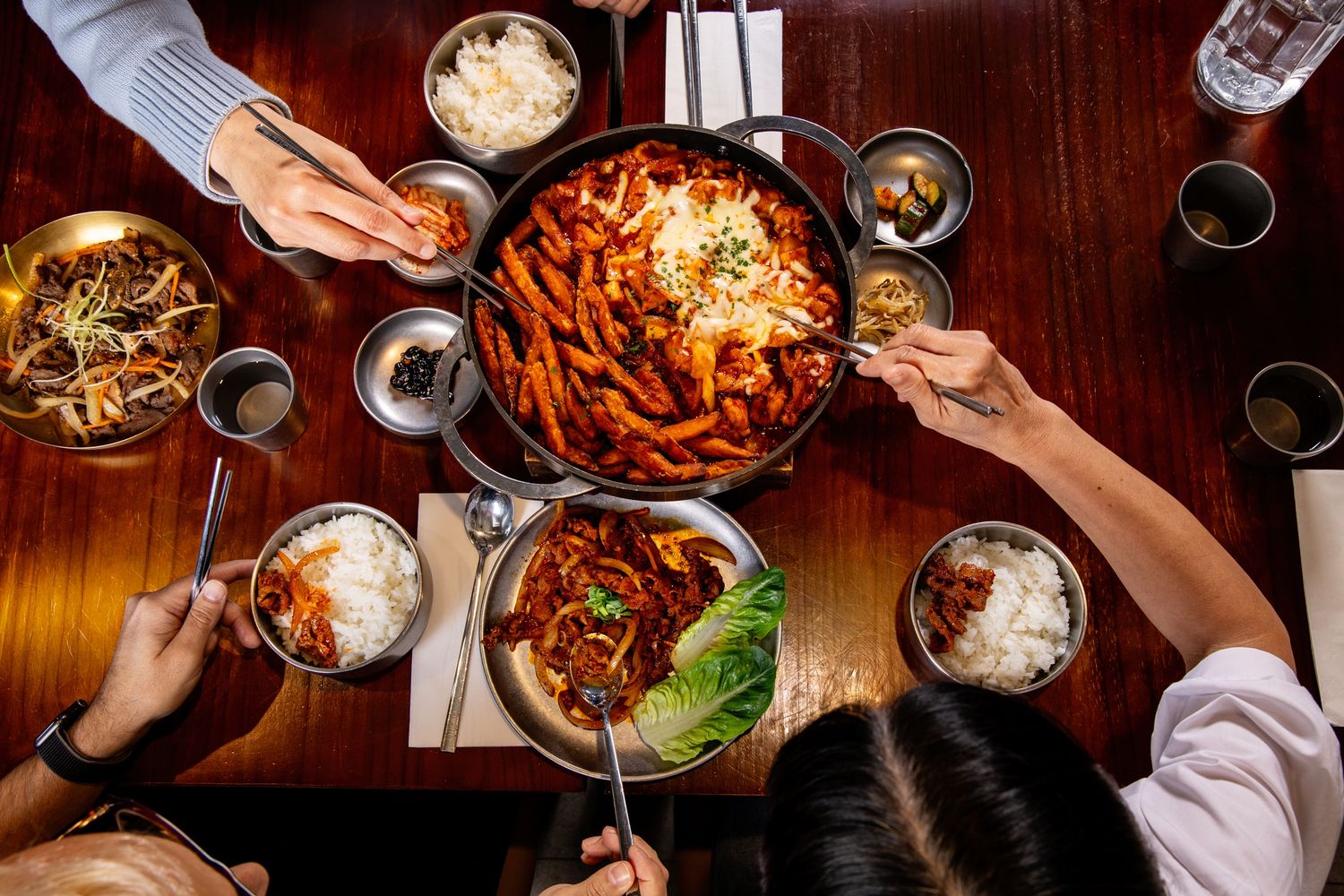 Overhead view of people sharing Korean tteokbokki and rice dishes around a wooden table with banchan sides.