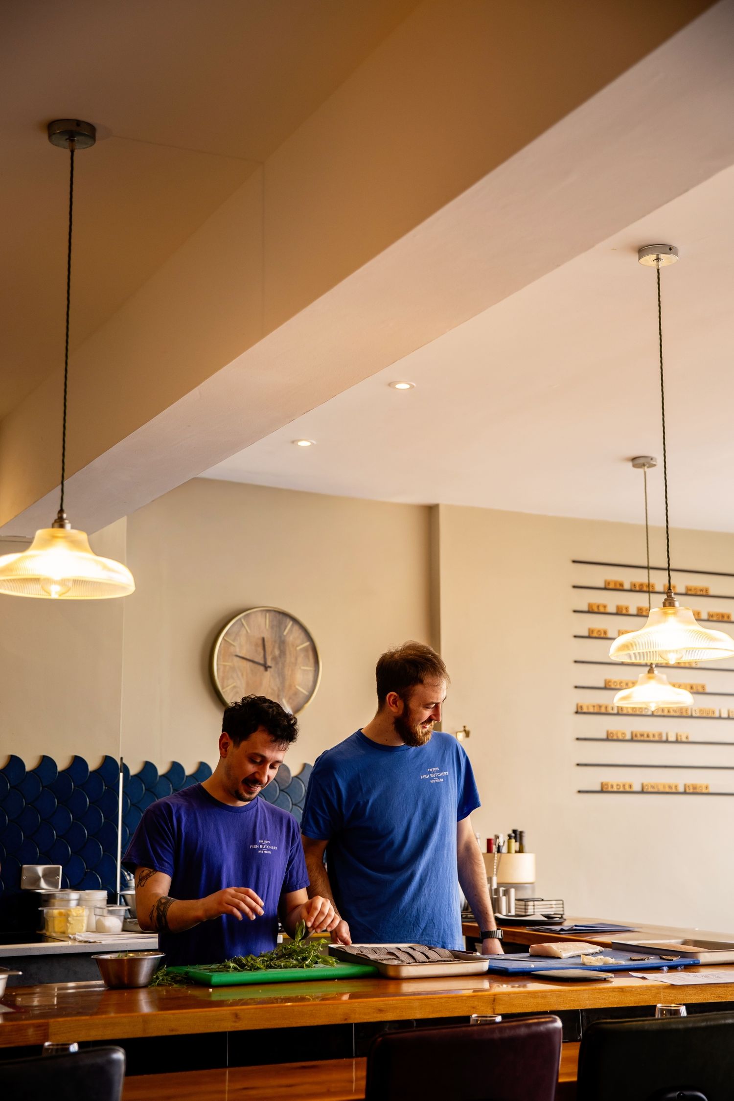 Two people work together behind a wooden bar counter in a restaurant with pendant lighting and shelving on the wall.