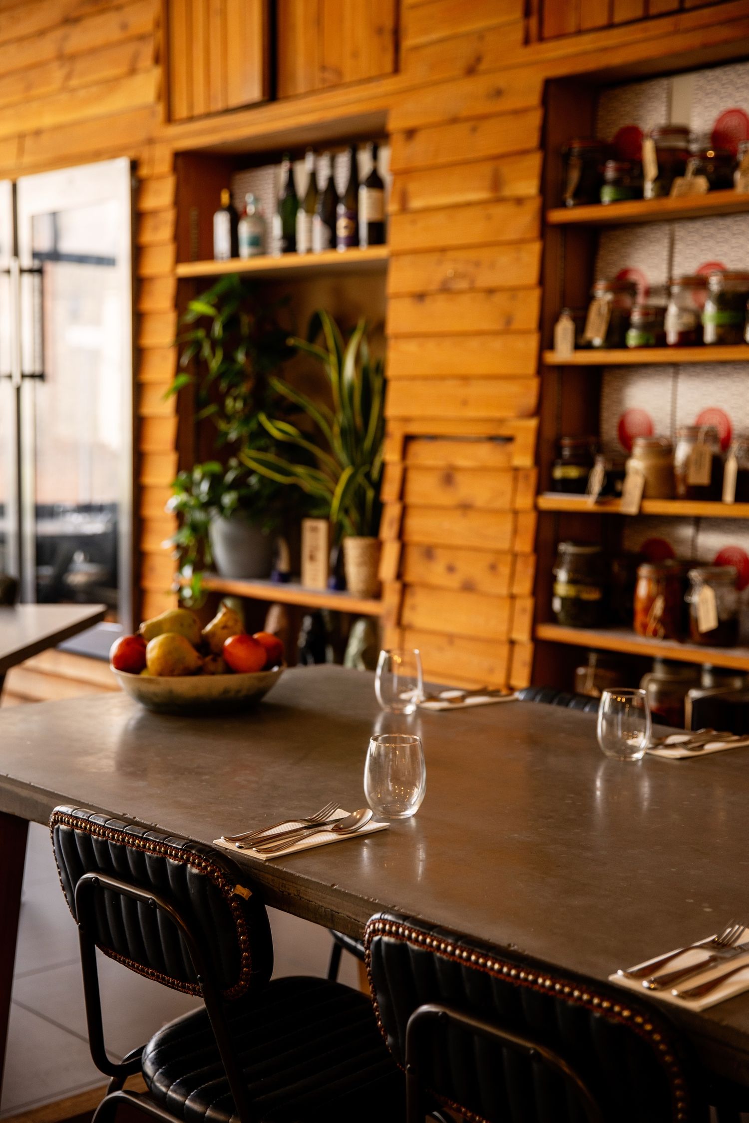 Rustic wooden restaurant interior with dark bar counter, shelves of bottles, and black leather bar stools.