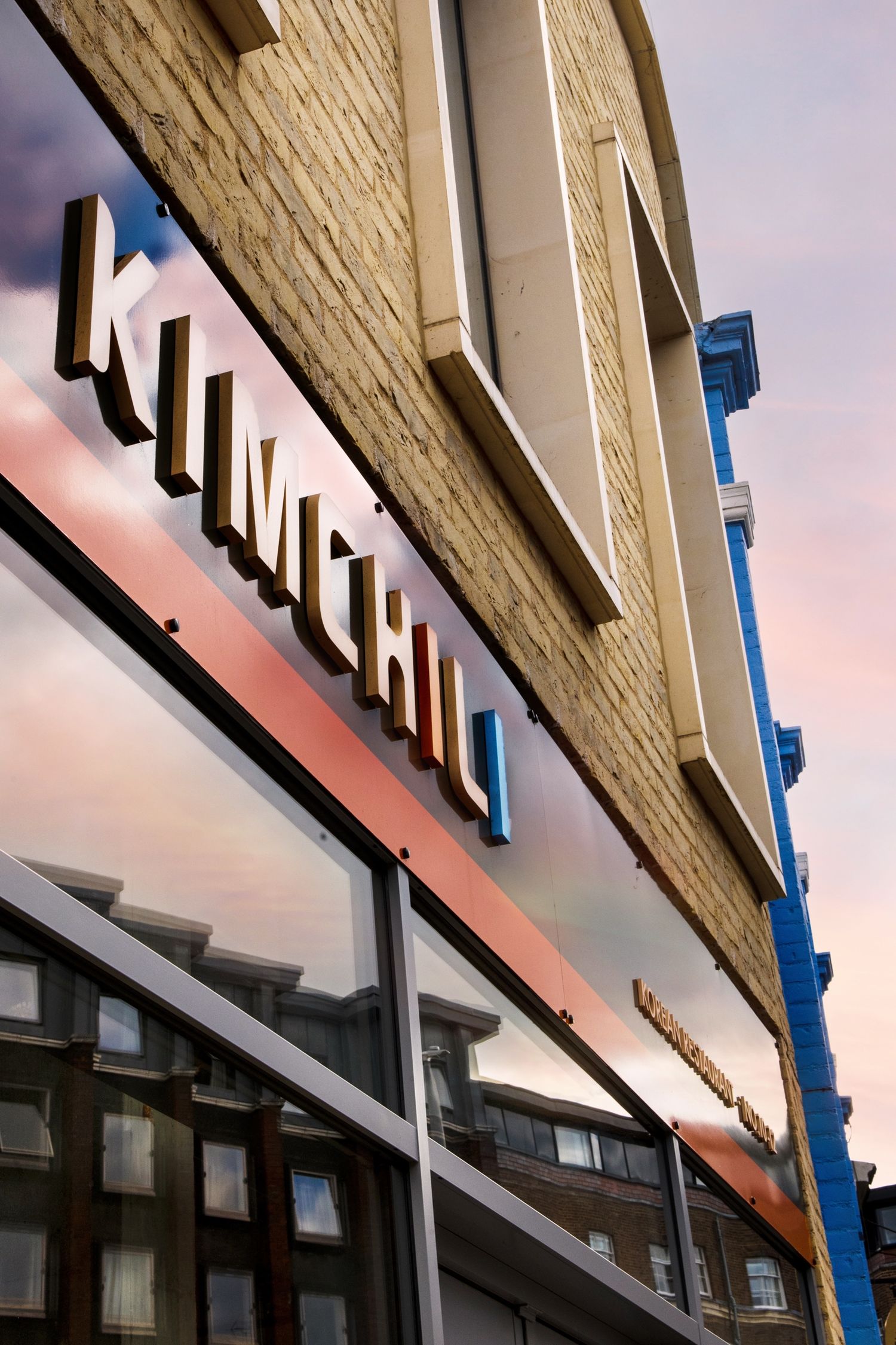 Exterior sign of Kimchi restaurant on a brick building facade reflecting colorful sunset sky in large windows.