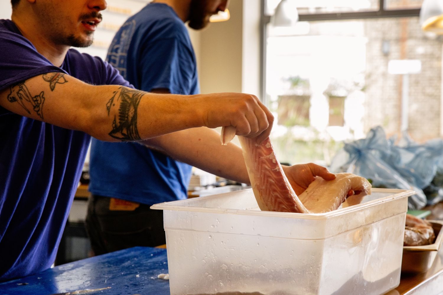 Person with tattooed arm seasoning food in a large white container in a kitchen setting.