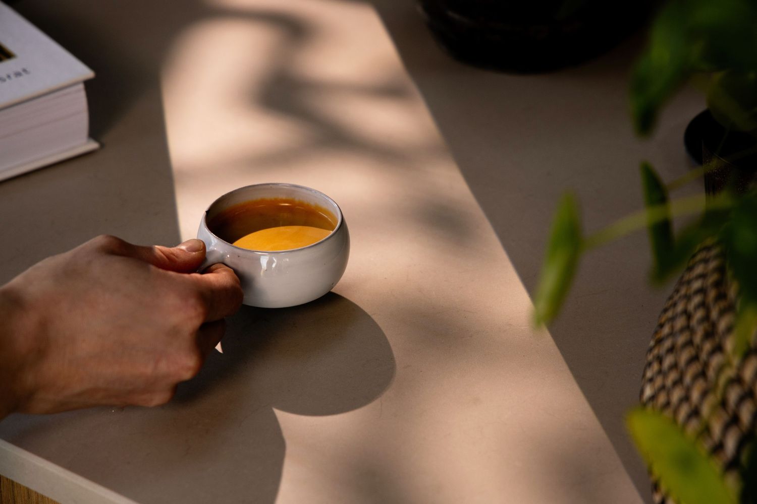 A hand holding a white ceramic cup of golden tea on a sunlit table with dappled shadows and green plants nearby.