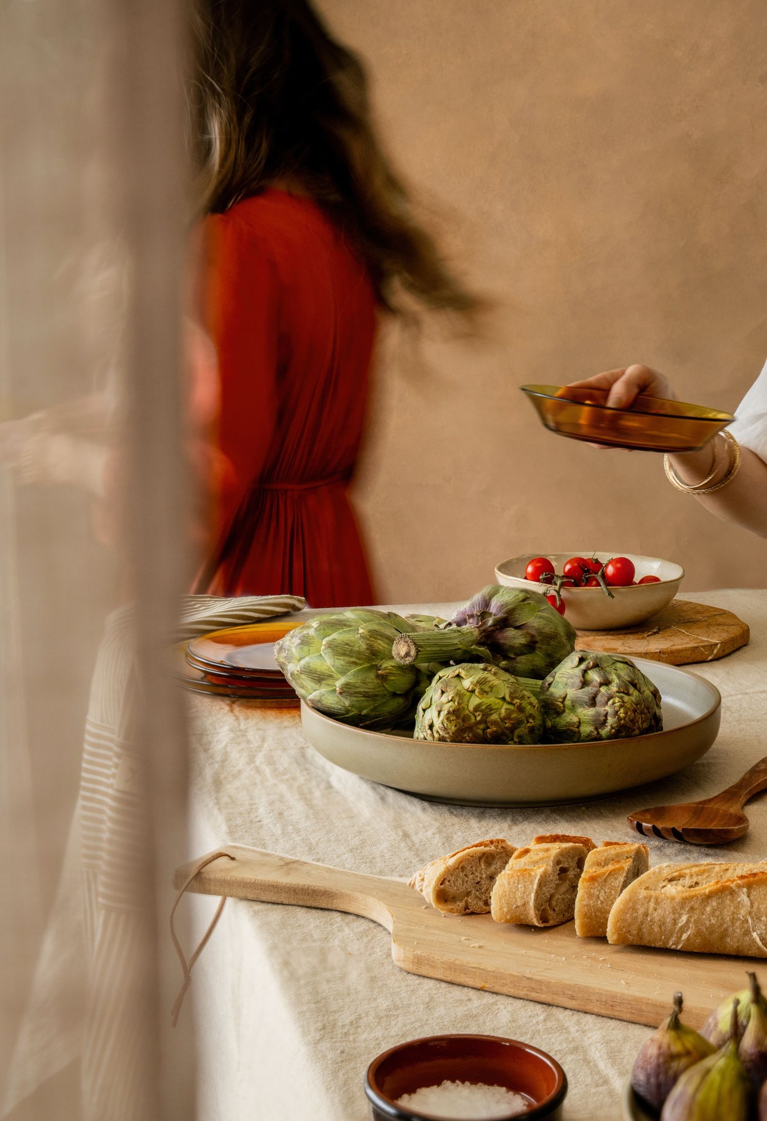 A rustic table spread with artichokes, bread, and tomatoes as a woman in a red dress stands nearby.