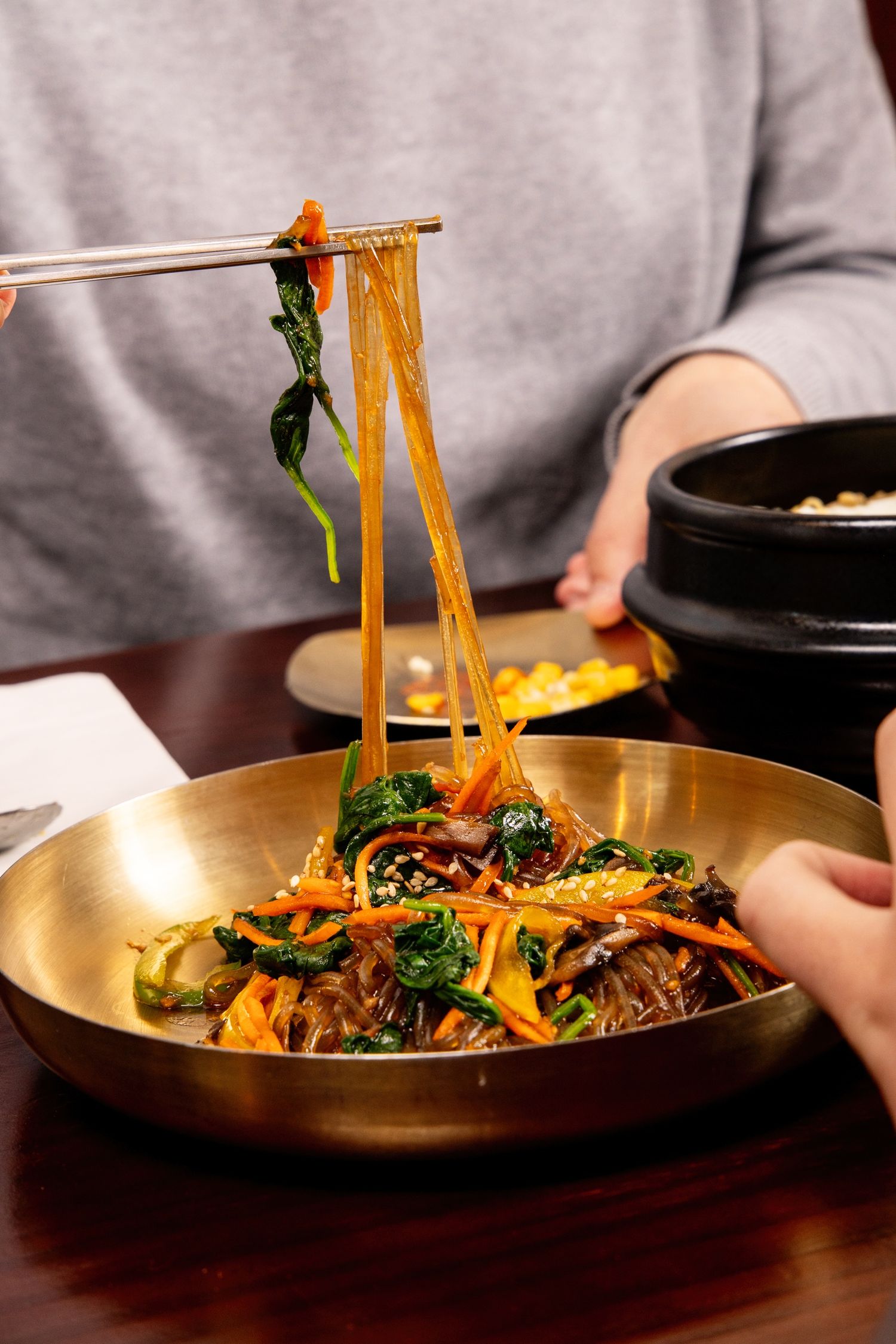 Chopsticks lifting glass noodles japchae with spinach and carrots from a golden bowl at a Korean restaurant.