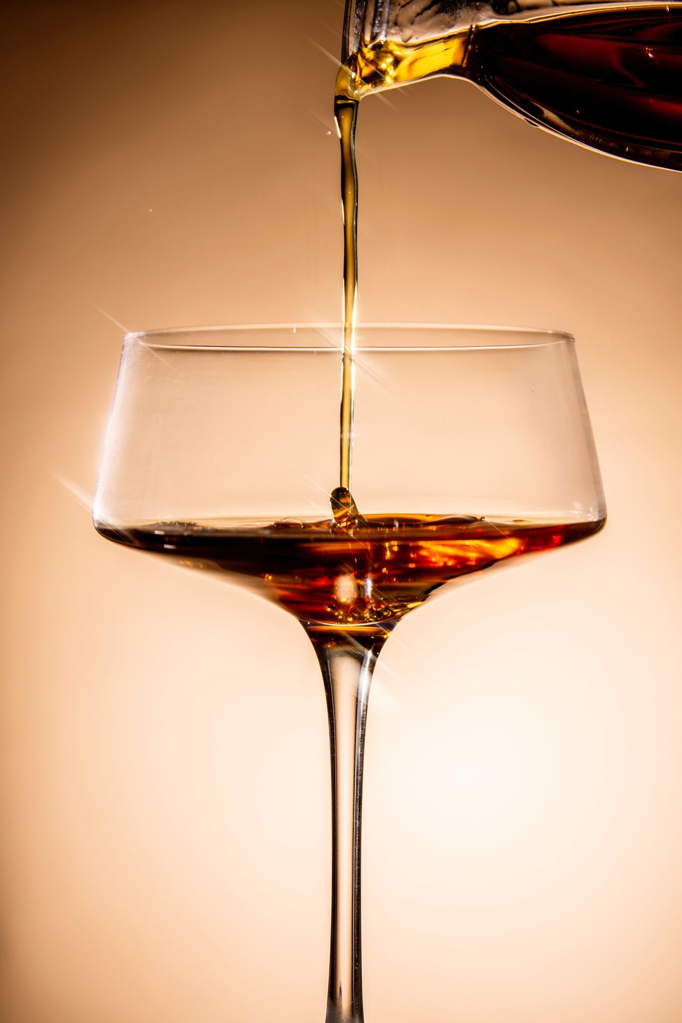 Amber liquid being poured into a champagne coupe glass against a warm beige background in a sequence of photos.