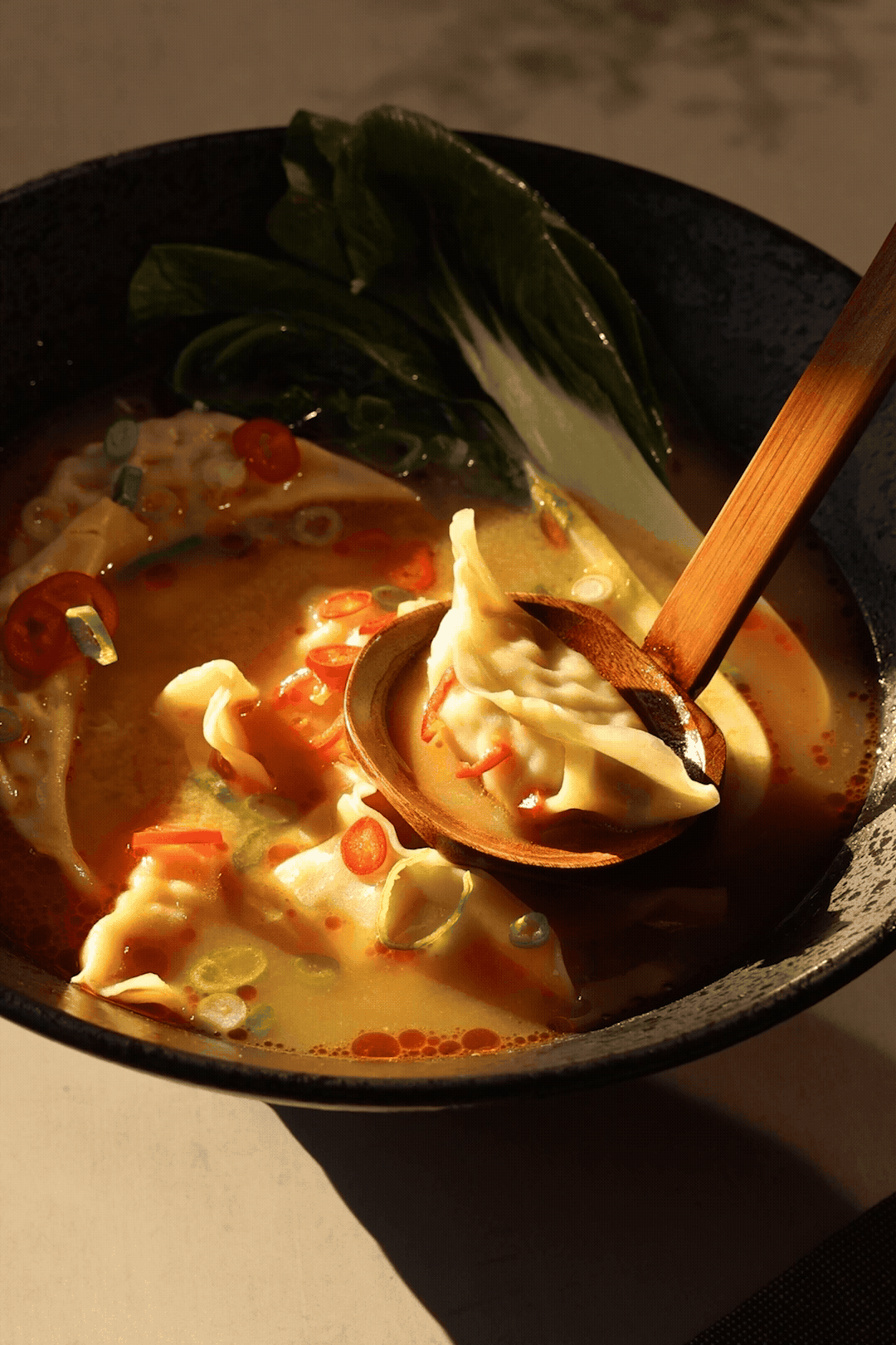 A steaming bowl of Asian dumpling soup with bok choy in a rich broth, served with a wooden spoon.