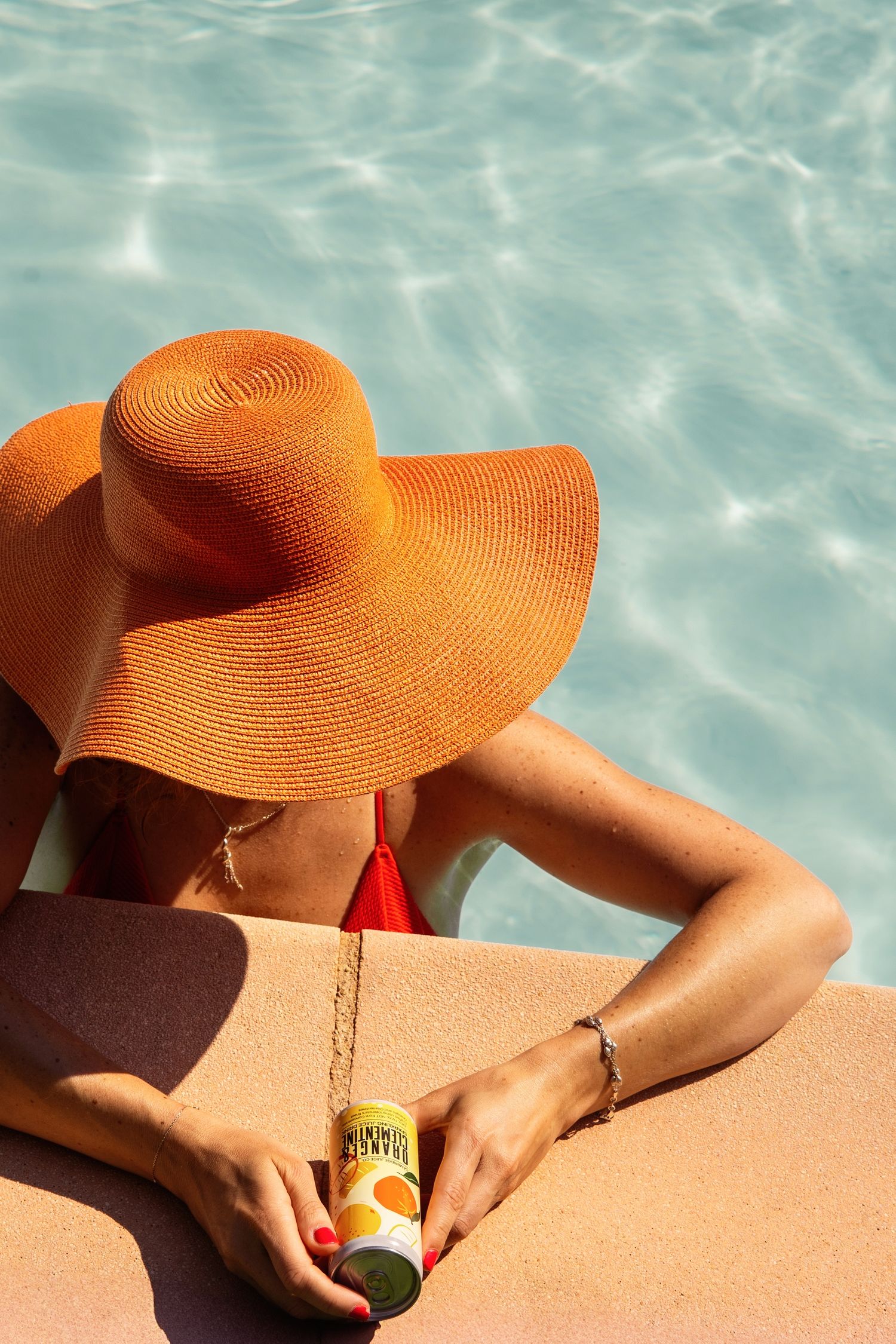 Orange sun hat and hands holding drink by pool with turquoise water.