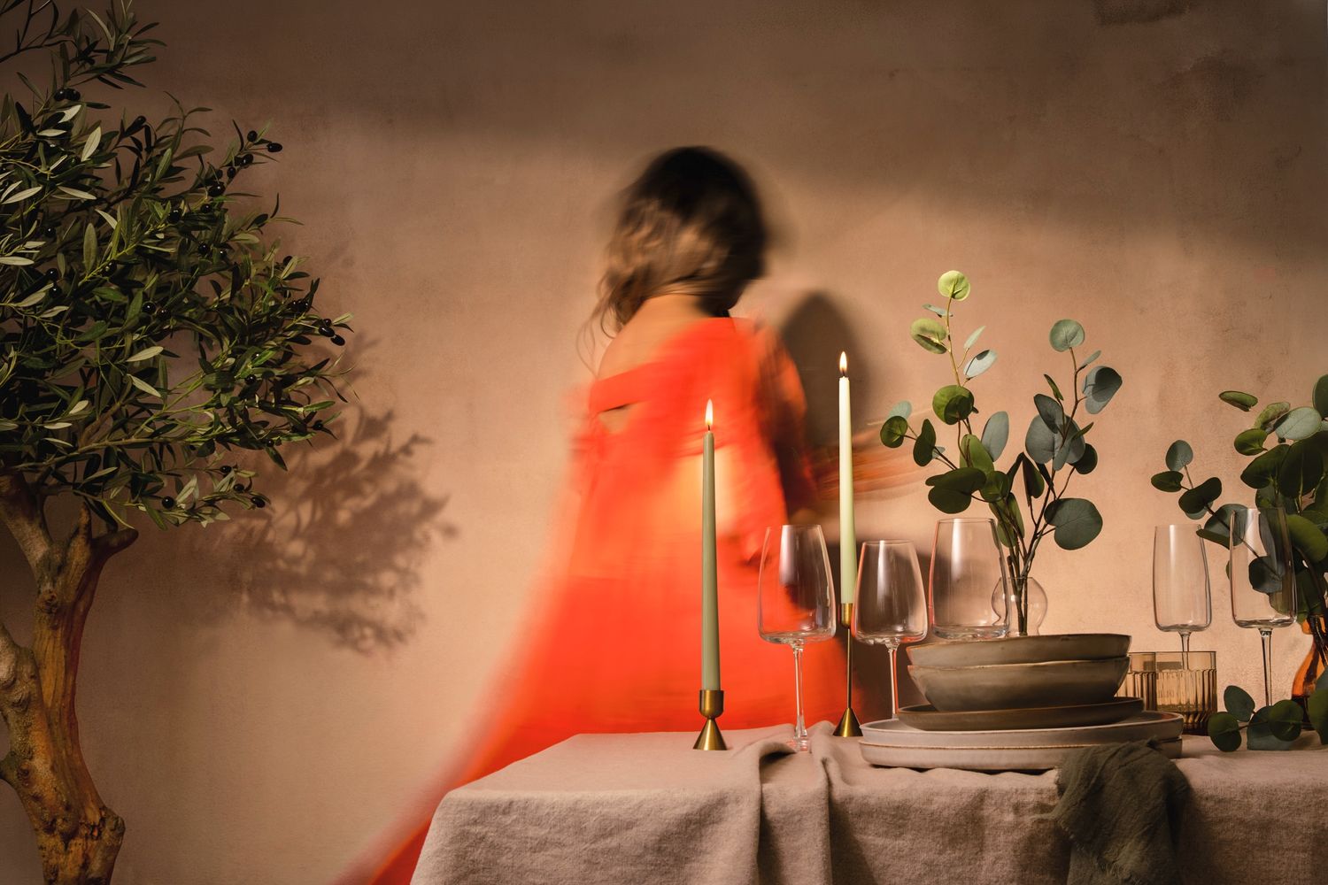 Blurred woman in orange dress standing by elegant table with candles, glassware, and greenery in warm candlelit setting.
