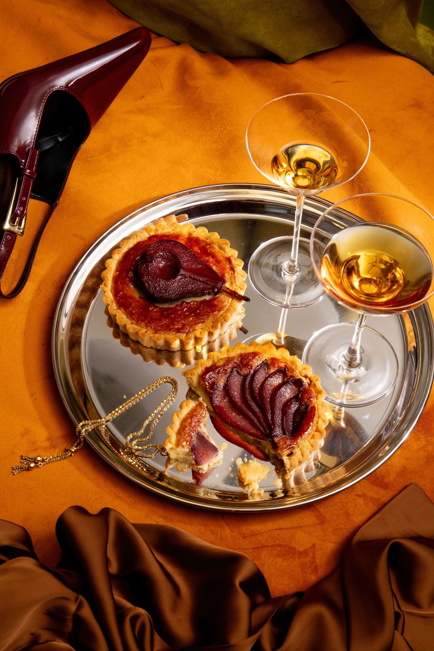 Elegant silver tray with pear tarts, two wine glasses, gold jewelry, on orange silk fabric with burgundy heels.