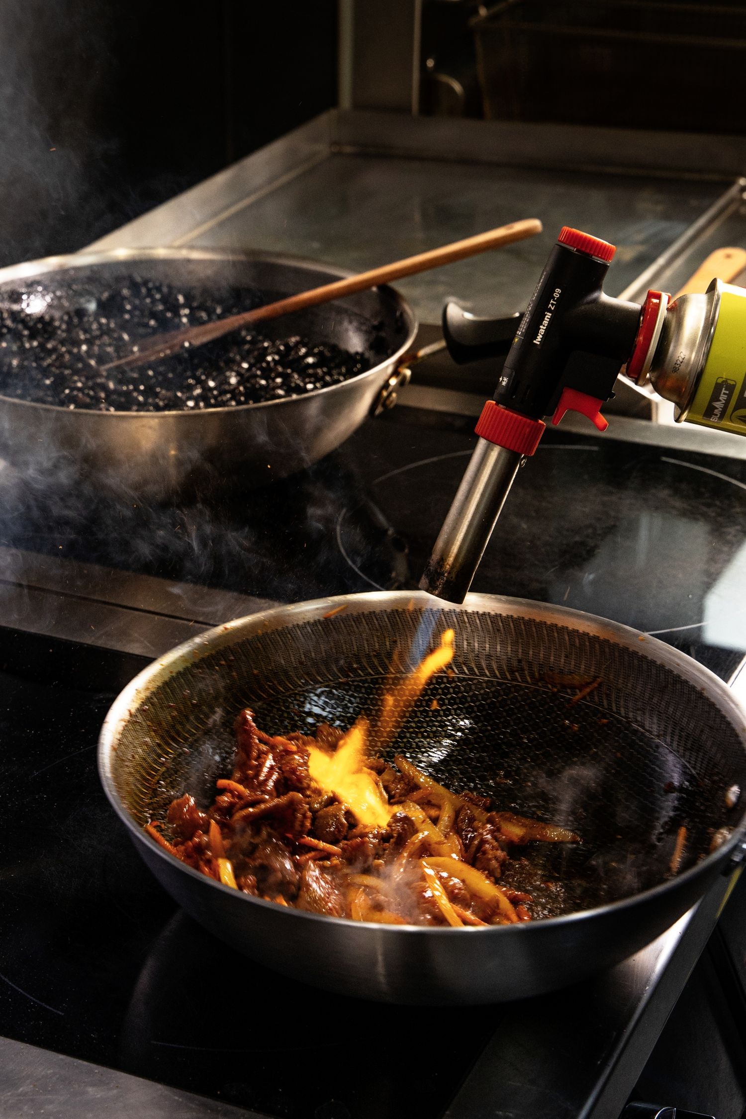 Chef using a blowtorch to flambé meat in a wok on an induction stovetop, with smoke rising dramatically.