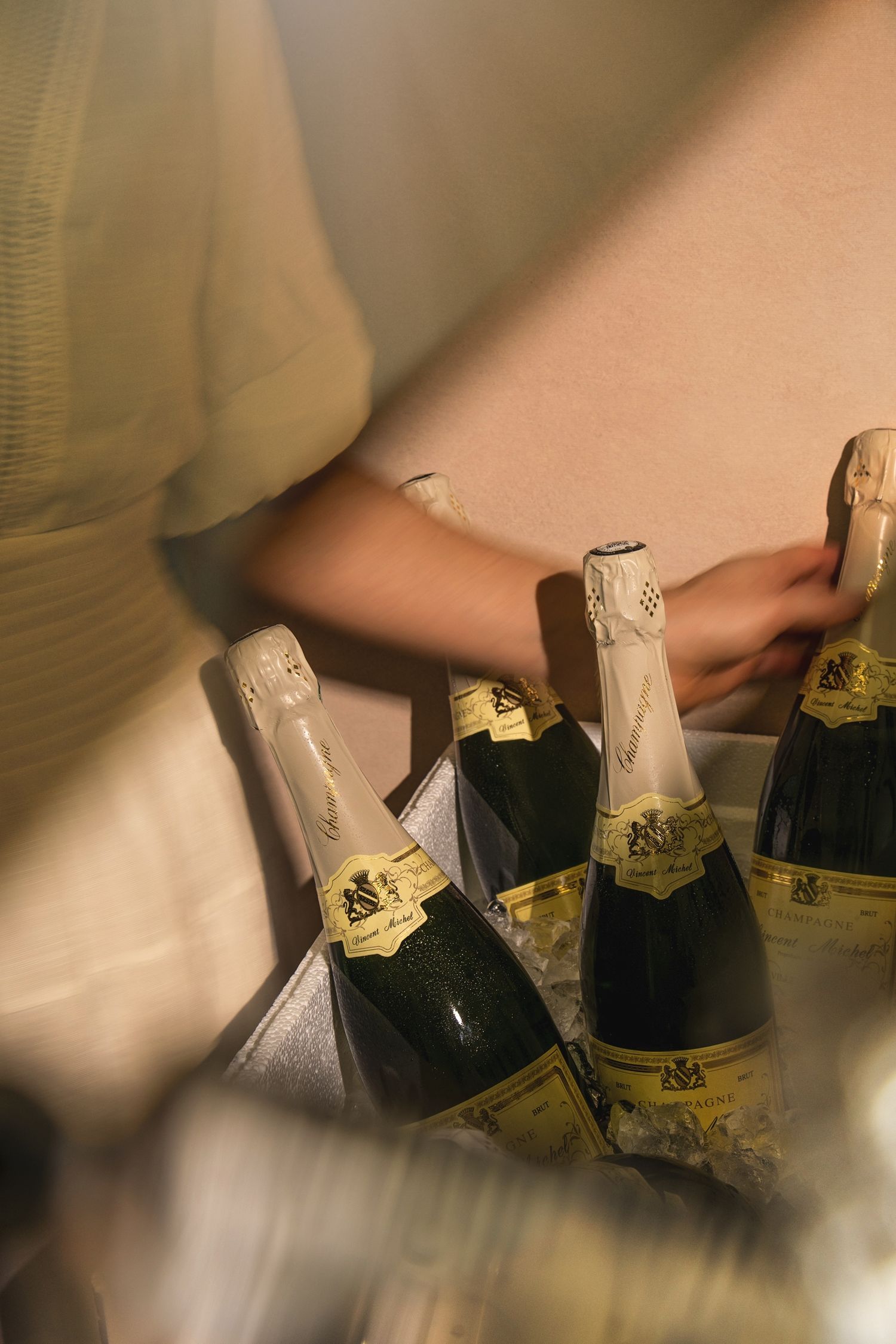 Person arranging multiple champagne bottles with gold labels on a surface in warm lighting.