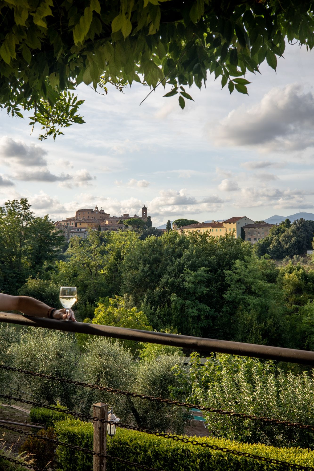 Scenic Tuscan landscape with hillside village and olive groves and glass of wine in hand under cloudy sky.