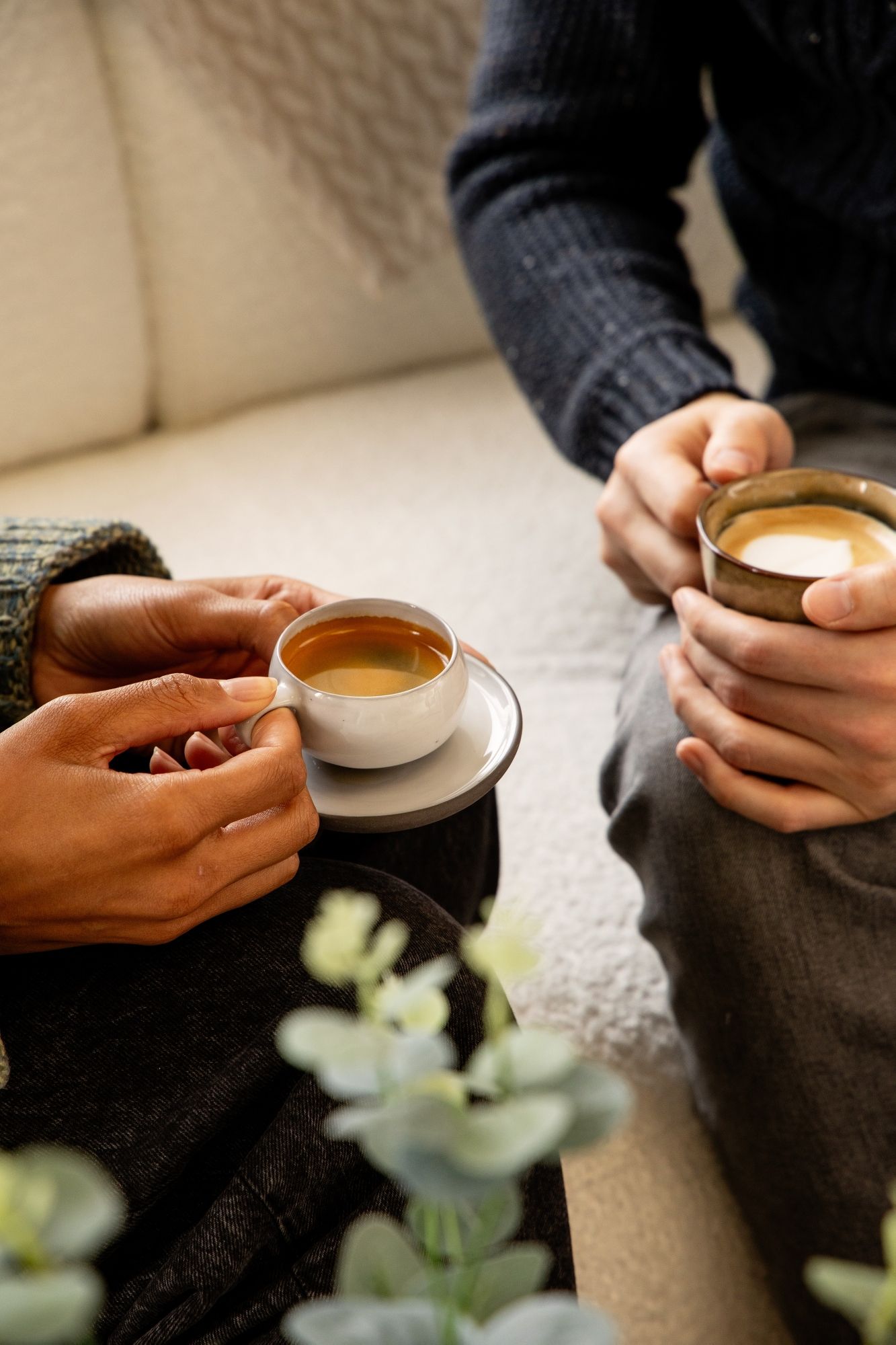 Hands holding coffee cups with latte art on a cosy sofa.