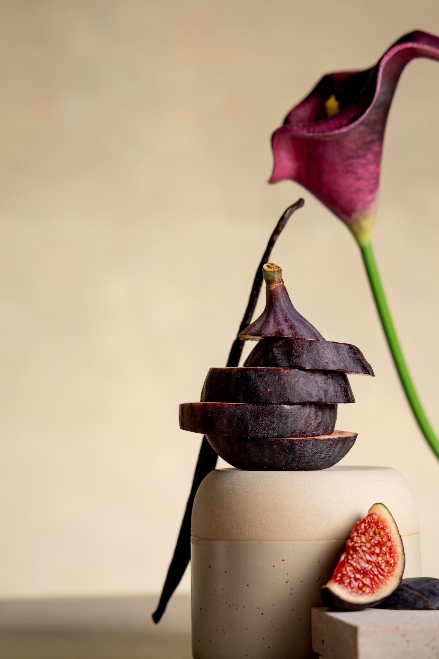 Sliced black figs stacked artistically beside a purple calla lily on a beige background with a halved fig showing red interior.