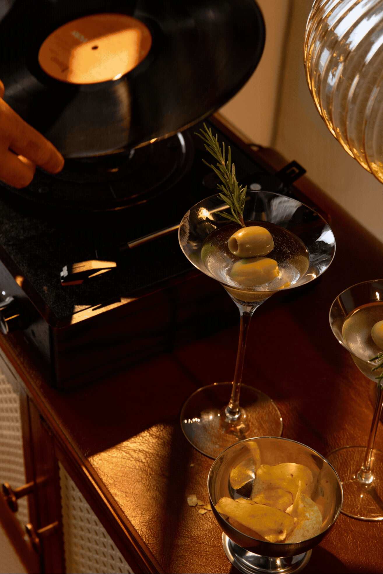 Elegant martini glass with olives and rosemary beside a vintage record player on a wooden bar cart.